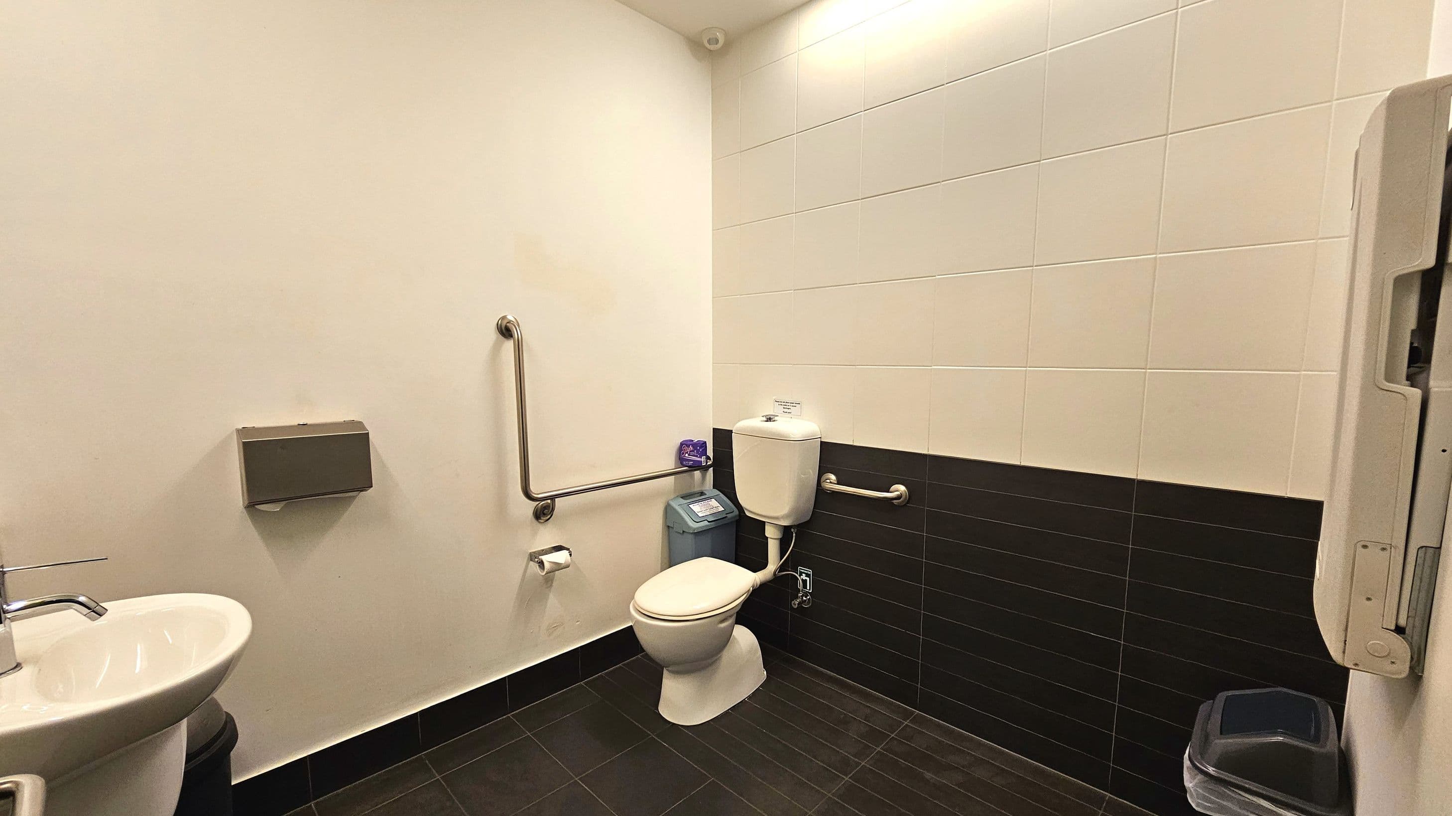 A public toilet with white tiled walls and dark tiled flooring. The toilet is positioned in the corner with a white cistern and a grab rail on the right. Another grab rail is mounted on the adjacent wall. A small bin is placed beside the toilet. A sink with a silver tap is on the left, with a paper towel dispenser above it. A baby changing station is folded against the wall on the right.