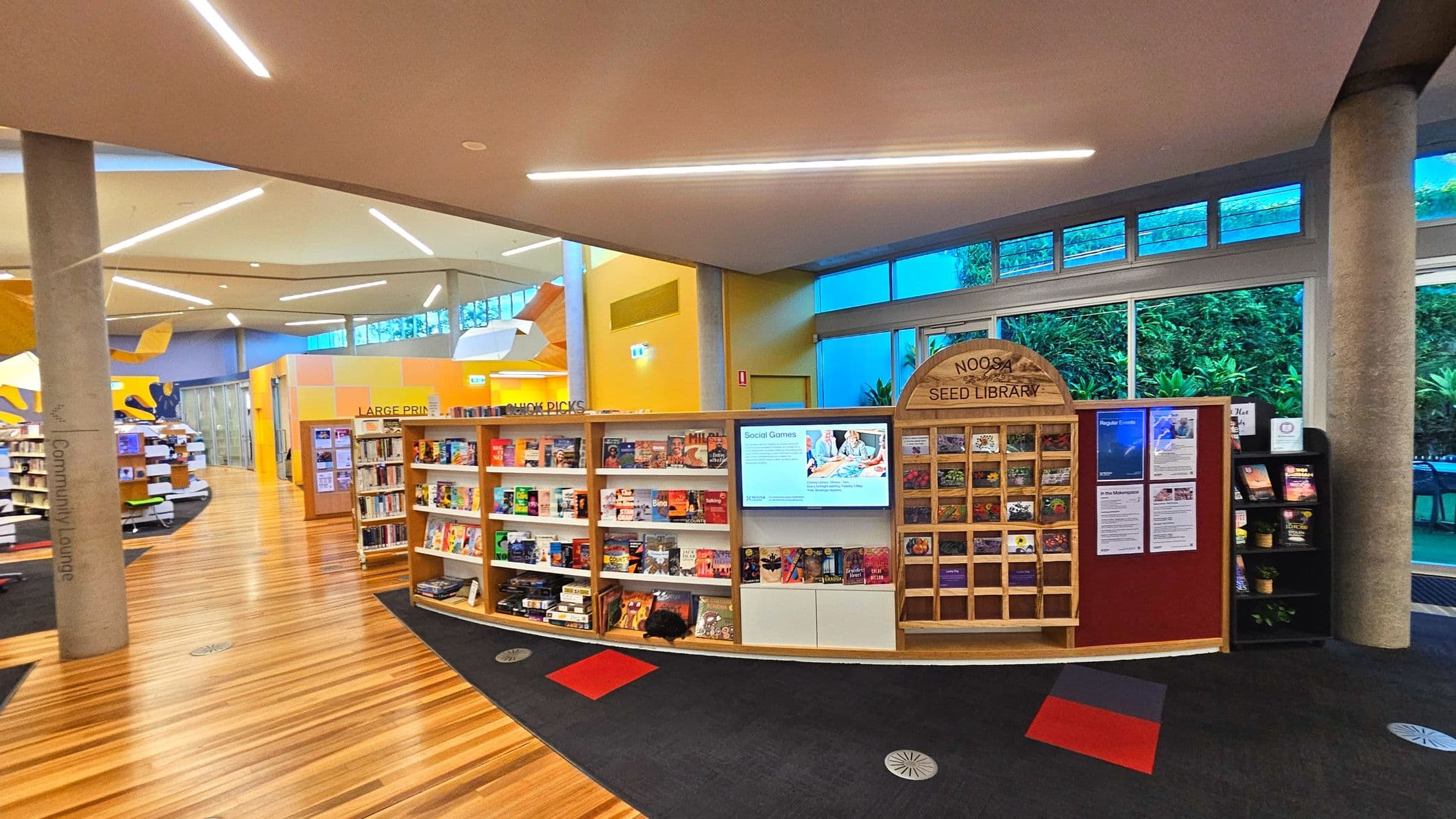 The image shows a modern library interior with a prominent wooden display labelled 'Noosa Seed Library' filled with seed packets. Adjacent is a bookshelf with various books and a digital screen displaying 'Social Games'. The floor transitions from light wood to dark carpet with red and grey accents. Large windows with greenery outside line the right wall, and the ceiling features angular lighting. Concrete pillars support the space.