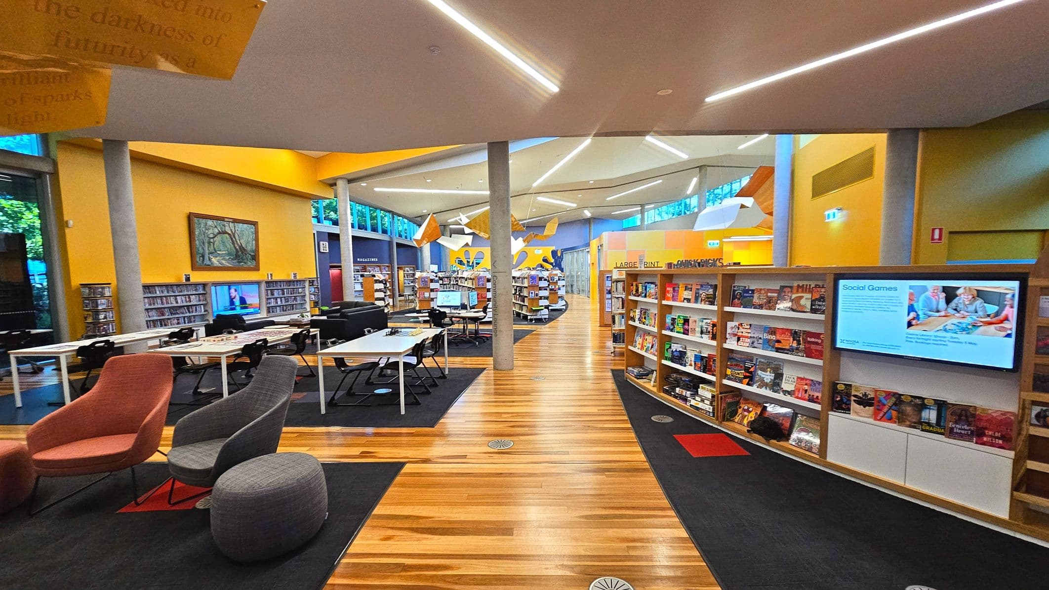 A modern library interior with a spacious layout featuring wooden floors and bright yellow walls. The space is divided by bookshelves and includes comfortable seating areas with colourful chairs and tables. A digital screen displays information on a wall. Large windows allow natural light, and the ceiling has geometric lighting fixtures. The area is open with visible bookshelves labelled for different sections.