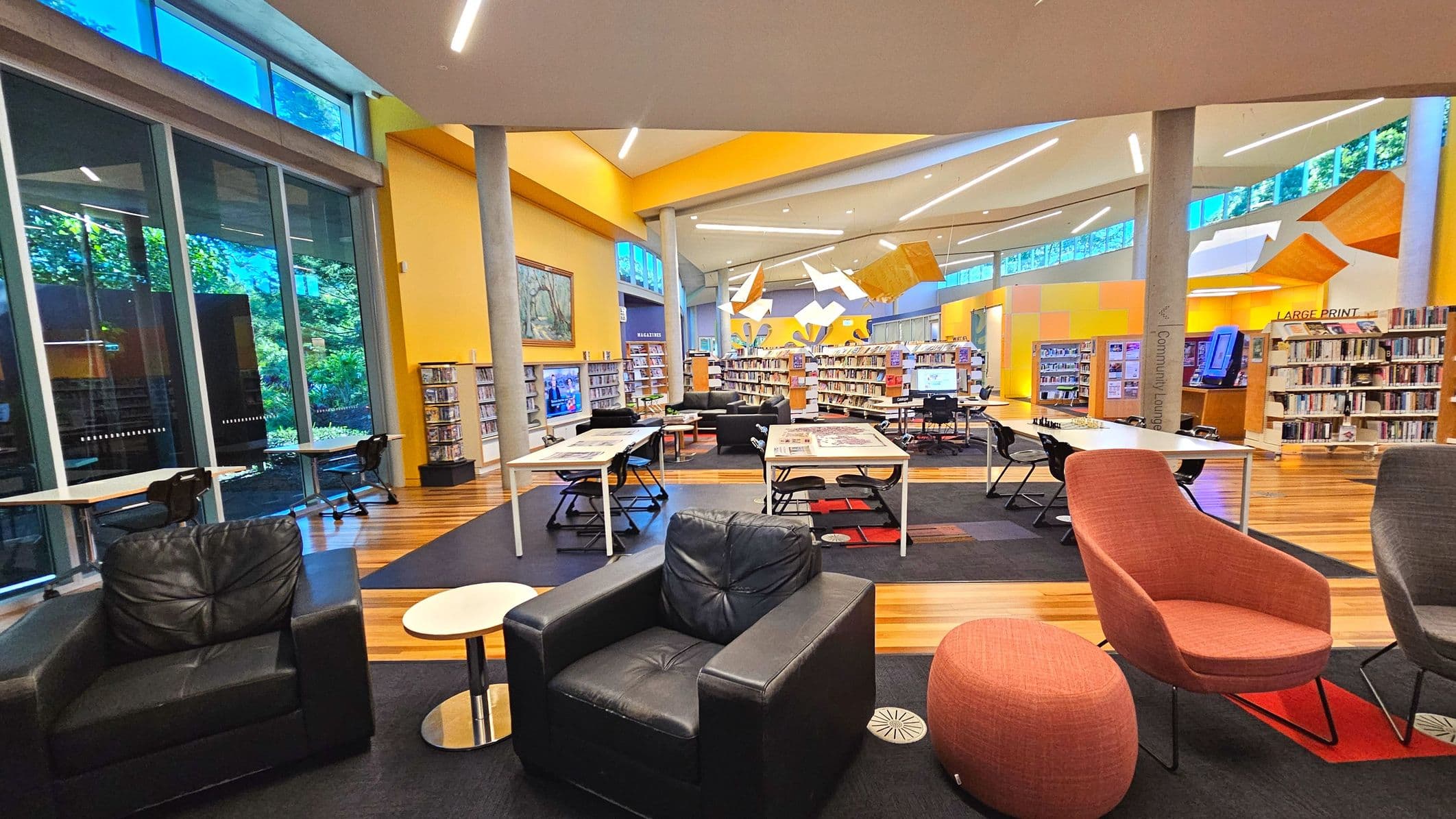 A spacious library interior with a modern design, featuring large glass windows on the left allowing natural light. The room has a high ceiling with angular light fixtures. Bookshelves line the walls, and there are several tables with chairs for reading. Comfortable armchairs and a small round table are in the foreground. The floor transitions from wood to carpet, and there are signs for 'Community Lounge' and 'Large Print'.