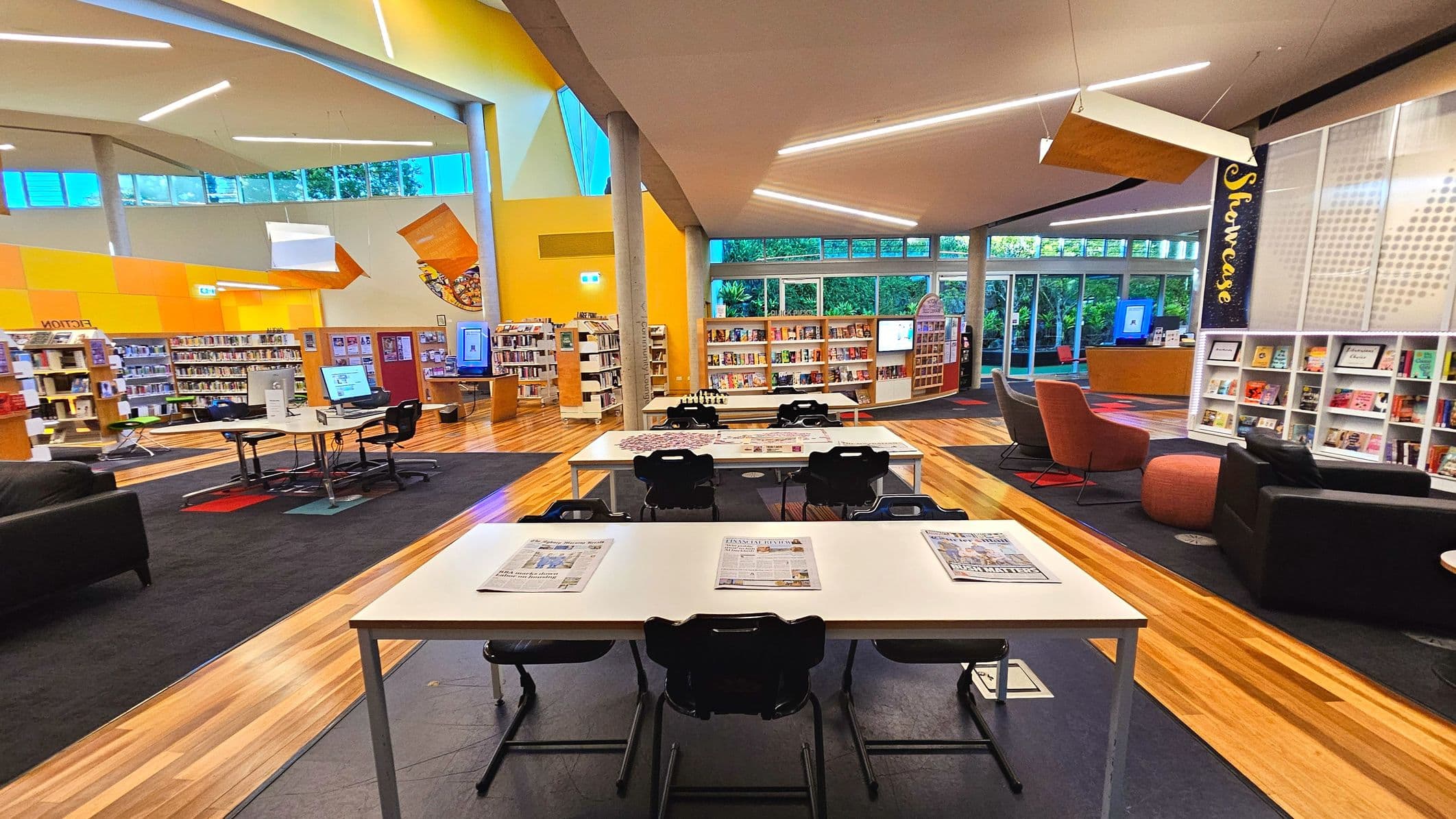 A modern library interior with wooden flooring and a mix of black and grey carpets. The space features white tables with black chairs, some with newspapers on them. Bookshelves line the walls, filled with books. A prominent 'Showcase' sign is visible on the right. Large windows allow natural light, and there are colourful chairs and a digital kiosk. The ceiling has angular designs with bright lighting.