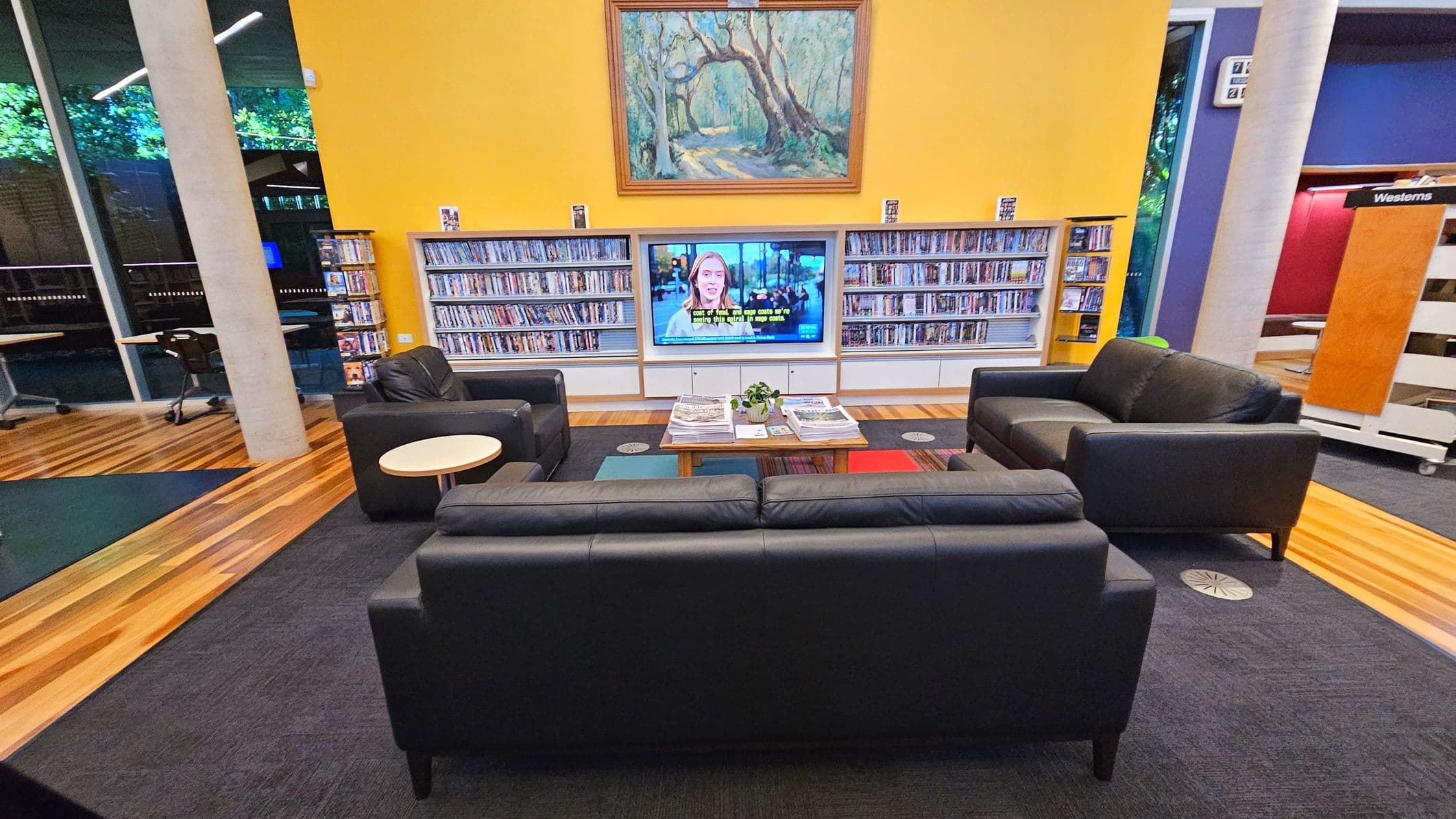 A library lounge area with a yellow wall featuring a large painting above a flat-screen TV. The TV is flanked by shelves filled with DVDs. In front of the TV, there are two black leather sofas and two armchairs arranged around a low table with magazines. The floor transitions from wooden planks to a dark carpet. Large windows and a concrete pillar are visible on the left, with a sign marked 'Westerns' on the right.