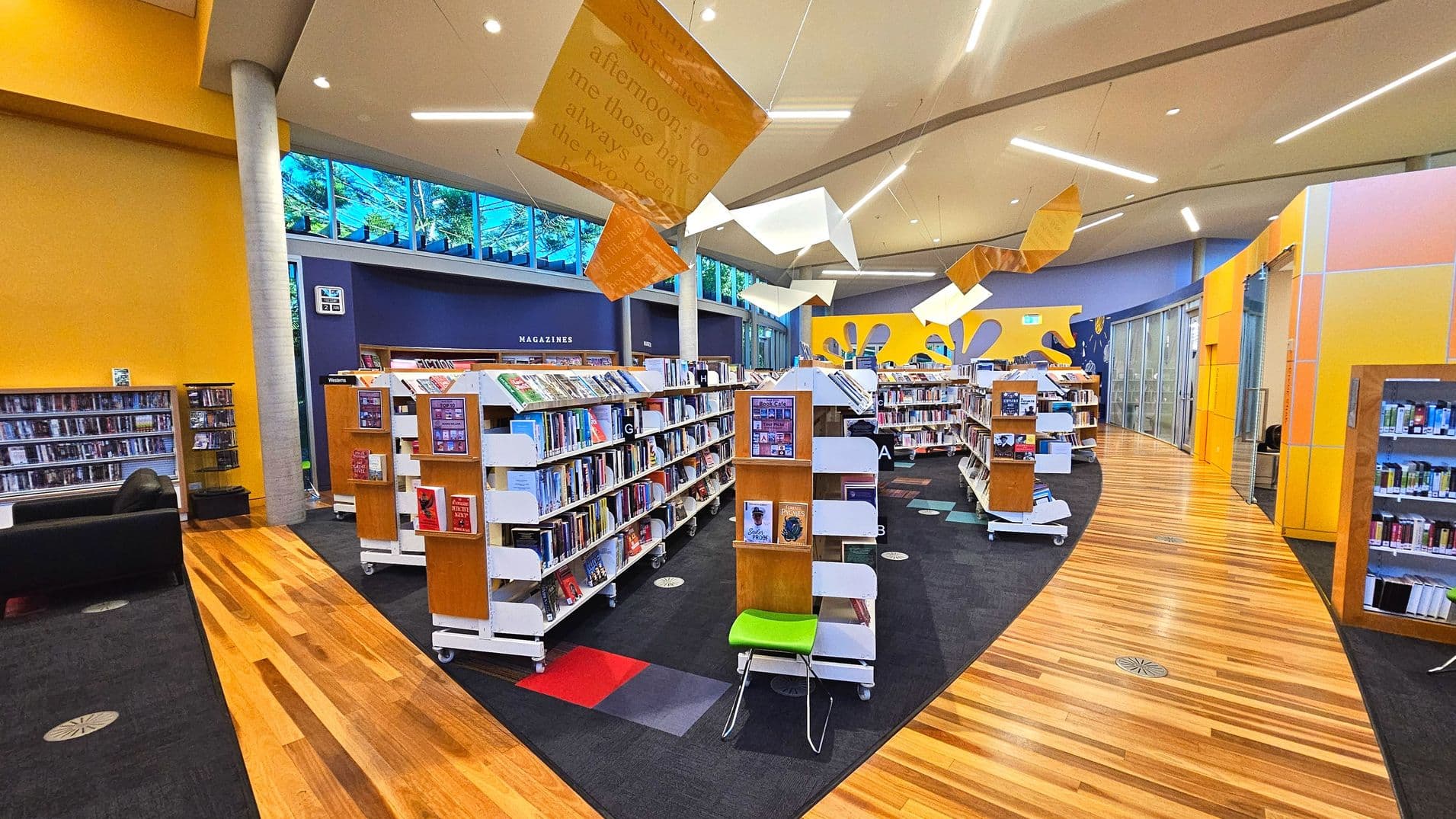 A modern library interior with vibrant yellow and blue walls. The space features several white bookcases filled with books, arranged in rows on a dark carpeted floor with colourful square patterns. A wooden pathway curves through the centre. Overhead, abstract geometric shapes hang from the ceiling. Large windows near the ceiling allow natural light in. A sign for 'MAGAZINES' is visible on a wall.