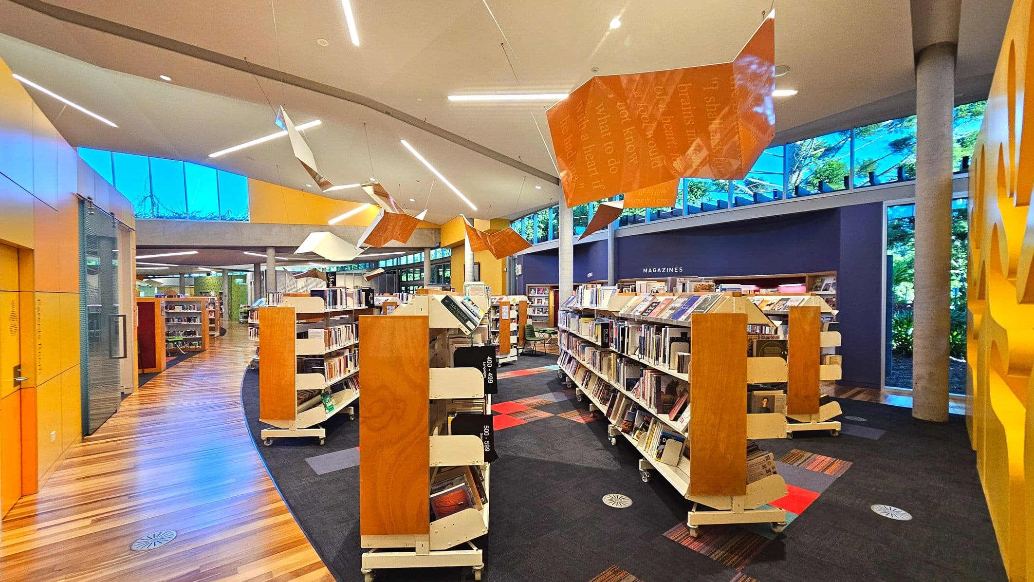 A modern library interior with curved wooden bookshelves on wheels, filled with books and magazines. The floor transitions from polished wood to patterned carpet. Bright orange geometric panels hang from the ceiling, which features recessed lighting. Large windows with blue-tinted glass line the upper walls, allowing natural light. A purple wall with 'MAGAZINES' signage is visible, and the space feels open with concrete columns.