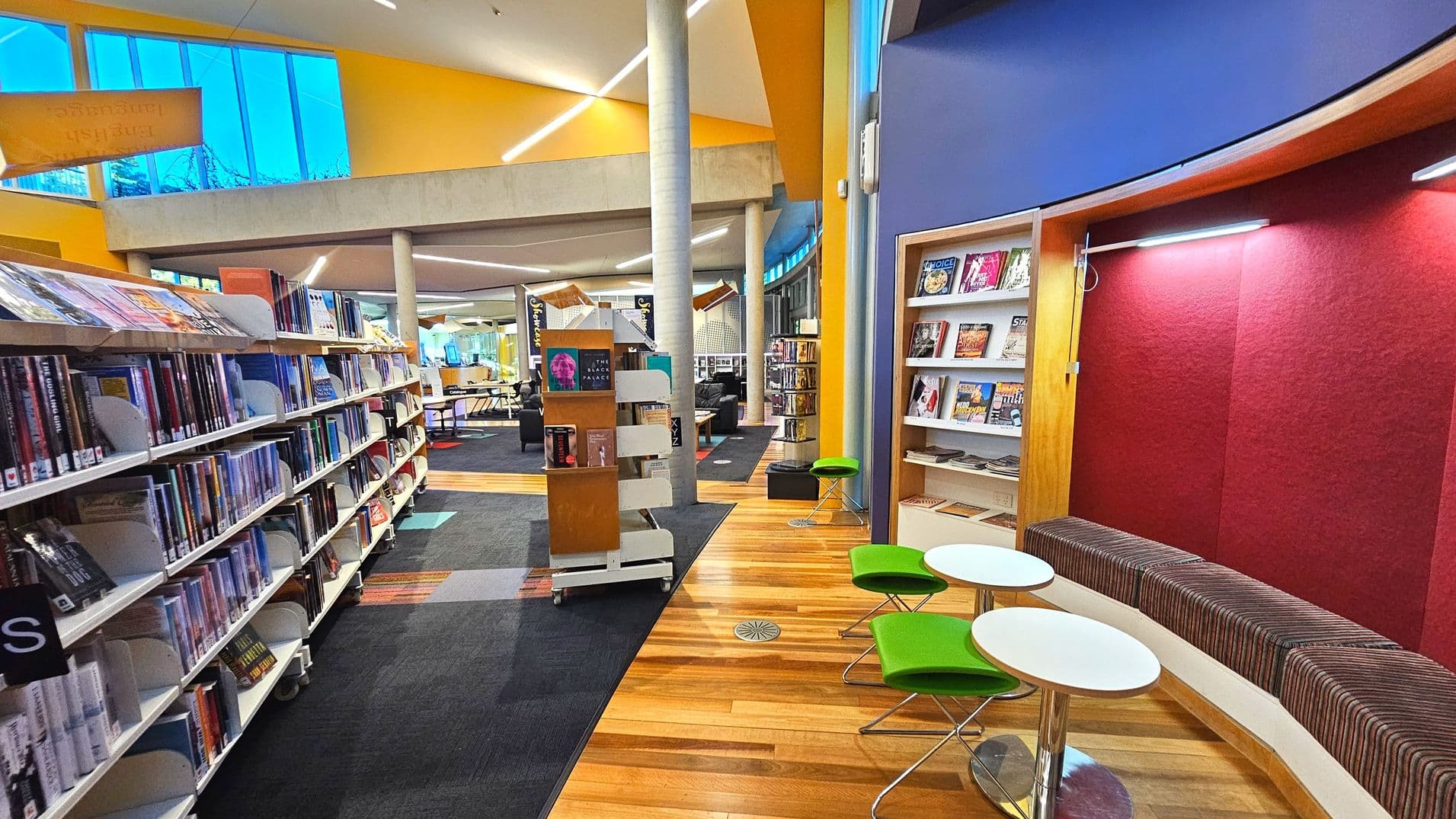 A modern library interior with tall bookshelves on the left filled with books. The floor transitions from dark carpet to light wooden planks. A curved seating area with striped cushions and small round tables with green stools is on the right. Tall columns support the high ceiling, and large windows allow natural light. A central display stand holds more books, and the space feels open and inviting.