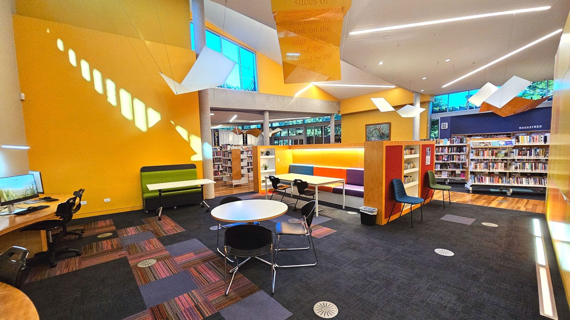 A modern library interior with vibrant yellow walls and large windows allowing natural light. The space features a mix of seating, including round tables with chairs, a green bench, and colourful booth seating. Bookshelves line the walls, with a section labelled 'MAGAZINES'. The floor has a mix of patterned carpet tiles and wood. Overhead, geometric light fixtures hang from the ceiling, adding a contemporary touch.