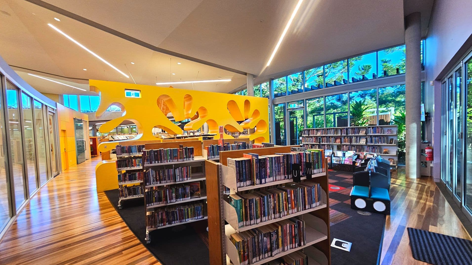 A modern library interior featuring a vibrant yellow partition with organic cut-out shapes, dividing the space. Bookshelves filled with books are arranged neatly on a wooden floor, transitioning to a carpeted area. Large glass windows line the right side, allowing natural light and views of greenery outside. The ceiling is high with sleek lighting fixtures, and a fire extinguisher is mounted on a column near the windows.