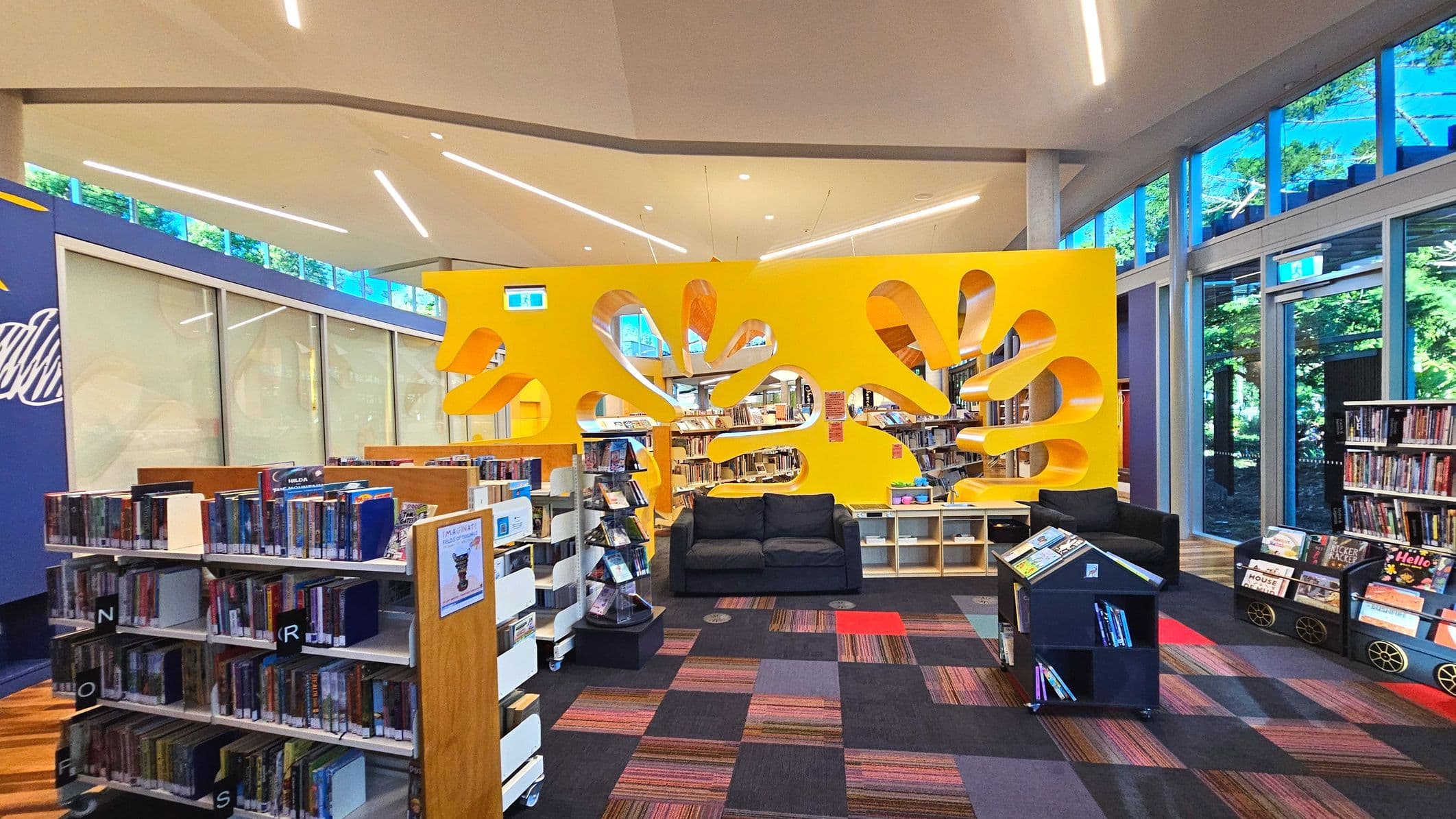 A modern library interior featuring a vibrant yellow partition with organic cut-out shapes, creating a playful design. Bookshelves filled with books line the space, and a black sofa sits in front of the partition. The floor is covered with a colourful, patterned carpet. Large windows on the right allow natural light to flood in, and the ceiling has sleek, linear lighting fixtures. A small book display stands in the foreground.