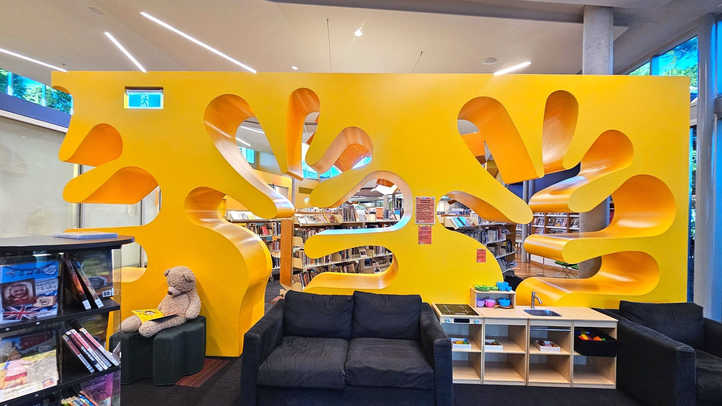 A vibrant library space featuring a large, bright yellow partition with organic, wavy cut-outs, creating a playful design. Behind it, bookshelves are visible, filled with books. In the foreground, a black sofa and a small play kitchen with a sink and shelves are present. A teddy bear sits on a small black stool holding a book. The area is well-lit with modern ceiling lights and large windows on the right.