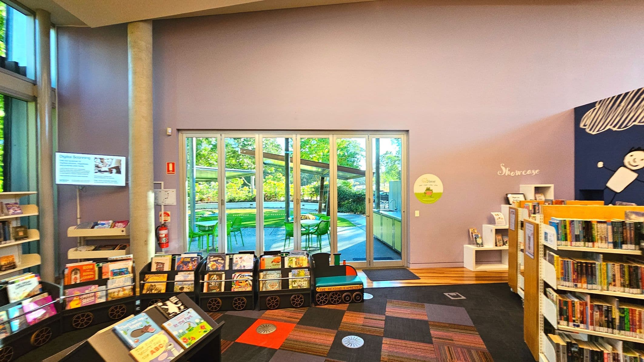 The image shows a library interior with a focus on a large glass door leading to an outdoor area with green chairs and tables. Inside, there are bookshelves filled with books, a digital screen on the left, and a playful train-shaped book display. The floor features a colourful carpet with geometric patterns. The walls are painted in soft purple, and a sign reading 'Showcase' is visible on the right.