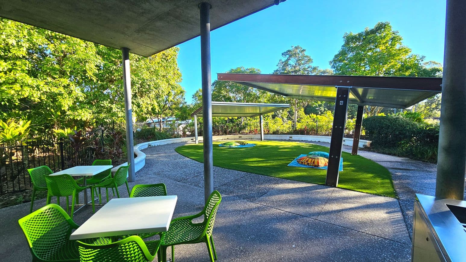 An outdoor area with a curved concrete path and a circular patch of artificial grass. Two modern metal shelters with transparent roofs provide shade over the grass. Bright green chairs and white tables are arranged on the path. The area is bordered by lush greenery and a black metal fence. The ground is a mix of pebbled concrete and artificial grass, creating a distinct surface transition.