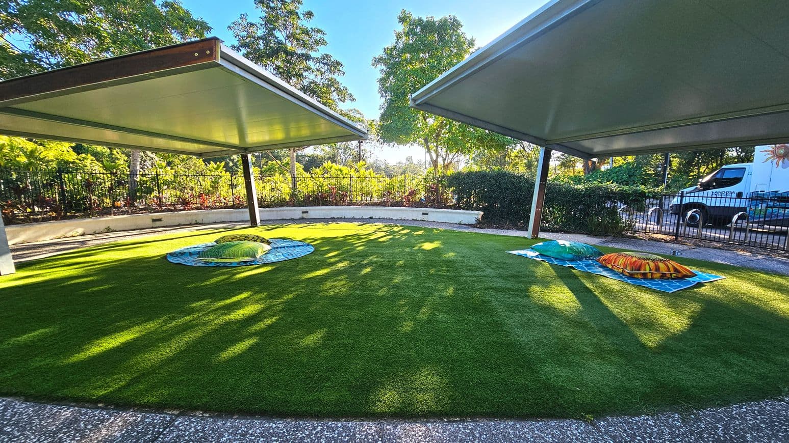 An outdoor area with two large, flat, metal canopies providing shade over a grassy lawn. The lawn features two colourful, turtle-shaped sculptures on circular mats. Surrounding the space is a low, white concrete wall with a black metal fence and lush greenery beyond. A paved path borders the grass, and a parked van is visible near the fence. The area feels open with ample natural light filtering through the trees.