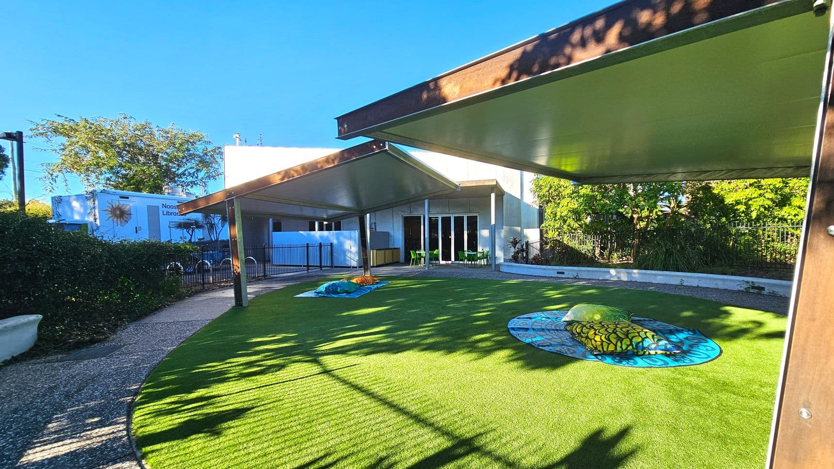 An outdoor area with artificial grass features two large, colourful play structures resembling insects. Overhead, three triangular shade sails provide cover. A curved gravel path runs alongside a hedge and leads to a building with glass doors and green chairs visible inside. A white truck with 'Noosa Library' branding is parked nearby. The space is bordered by a low fence and lush greenery.