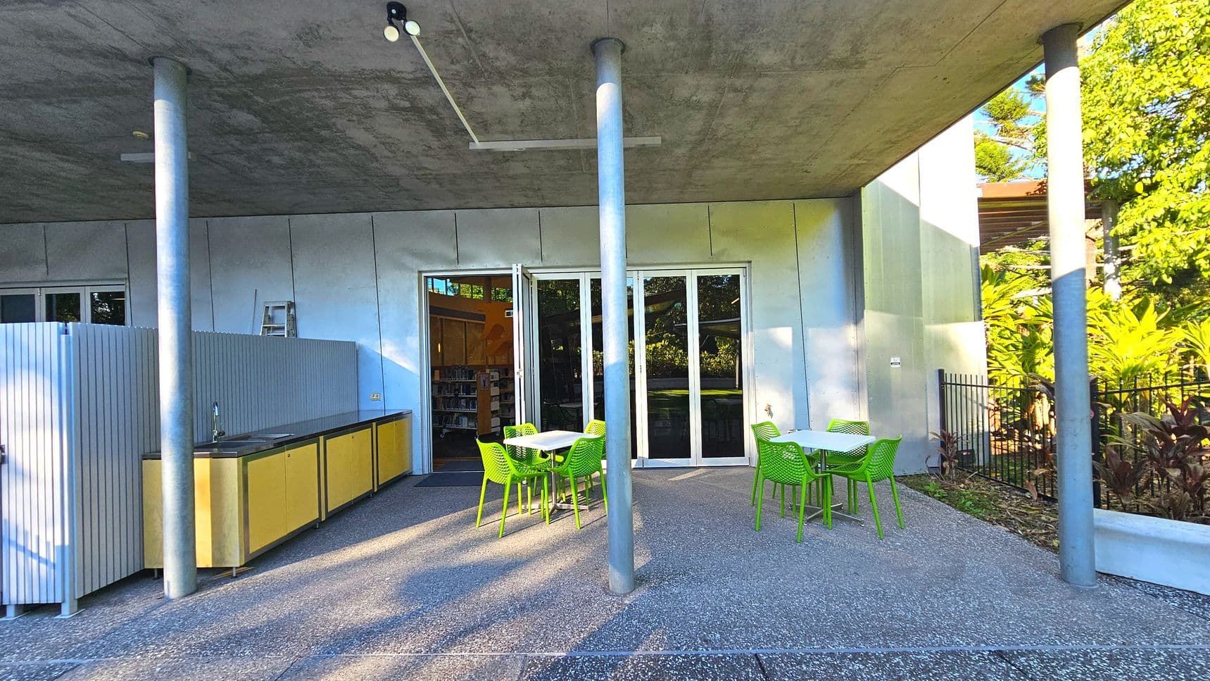 An outdoor area with a concrete ceiling supported by metal poles. The space features a kitchenette with yellow cabinets and a sink on the left, partially enclosed by a grey panel. In the centre, there are green chairs around white tables on a speckled concrete floor. Glass doors lead inside to a room with bookshelves. A black metal fence and lush greenery are visible on the right.