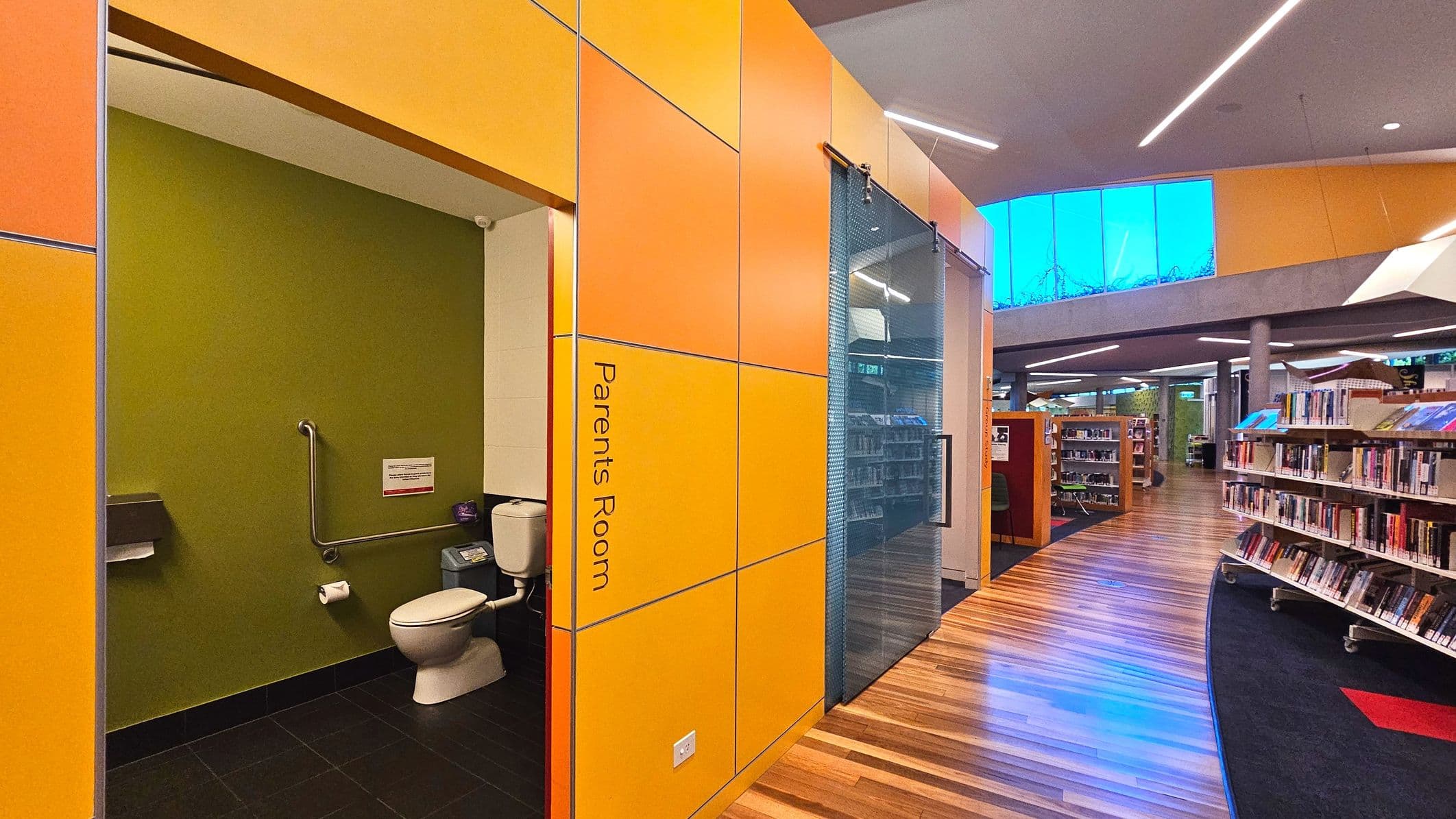 The image shows a library interior with a prominent orange-tiled wall labelled 'Parents Room' on the left. Inside, a toilet and grab bar are visible against a green wall. The library features wooden flooring transitioning to carpet, with bookshelves lining a curved path. Overhead, modern lighting and large windows provide illumination. The space feels open with visible concrete columns and a high ceiling.
