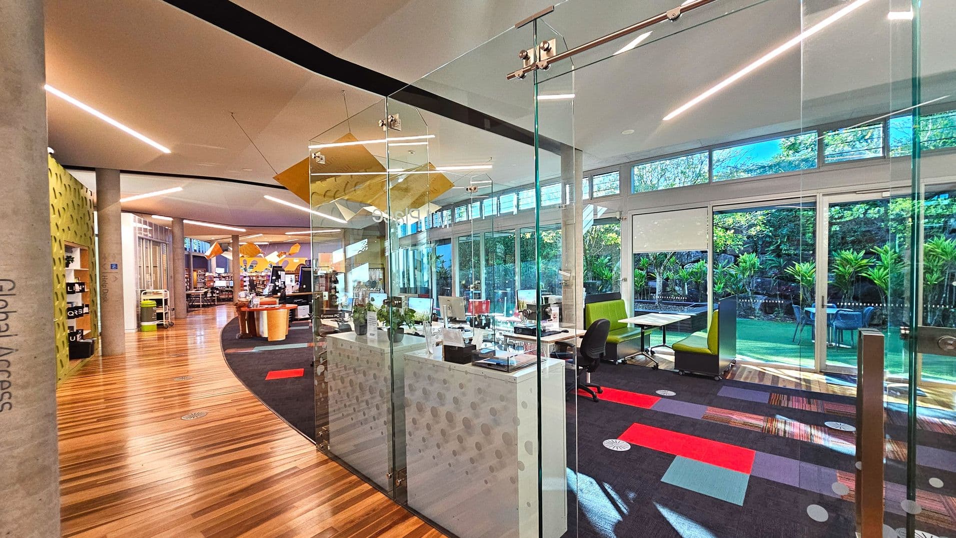 The image shows a modern interior space with a curved wooden floor leading into a library area. On the right, a glass-walled office with desks and chairs is visible, featuring a patterned carpet with red and purple accents. Large windows provide a view of lush greenery outside. The ceiling is high with linear lights, and there are cylindrical columns throughout. A sign on the wall reads 'Global Access'.