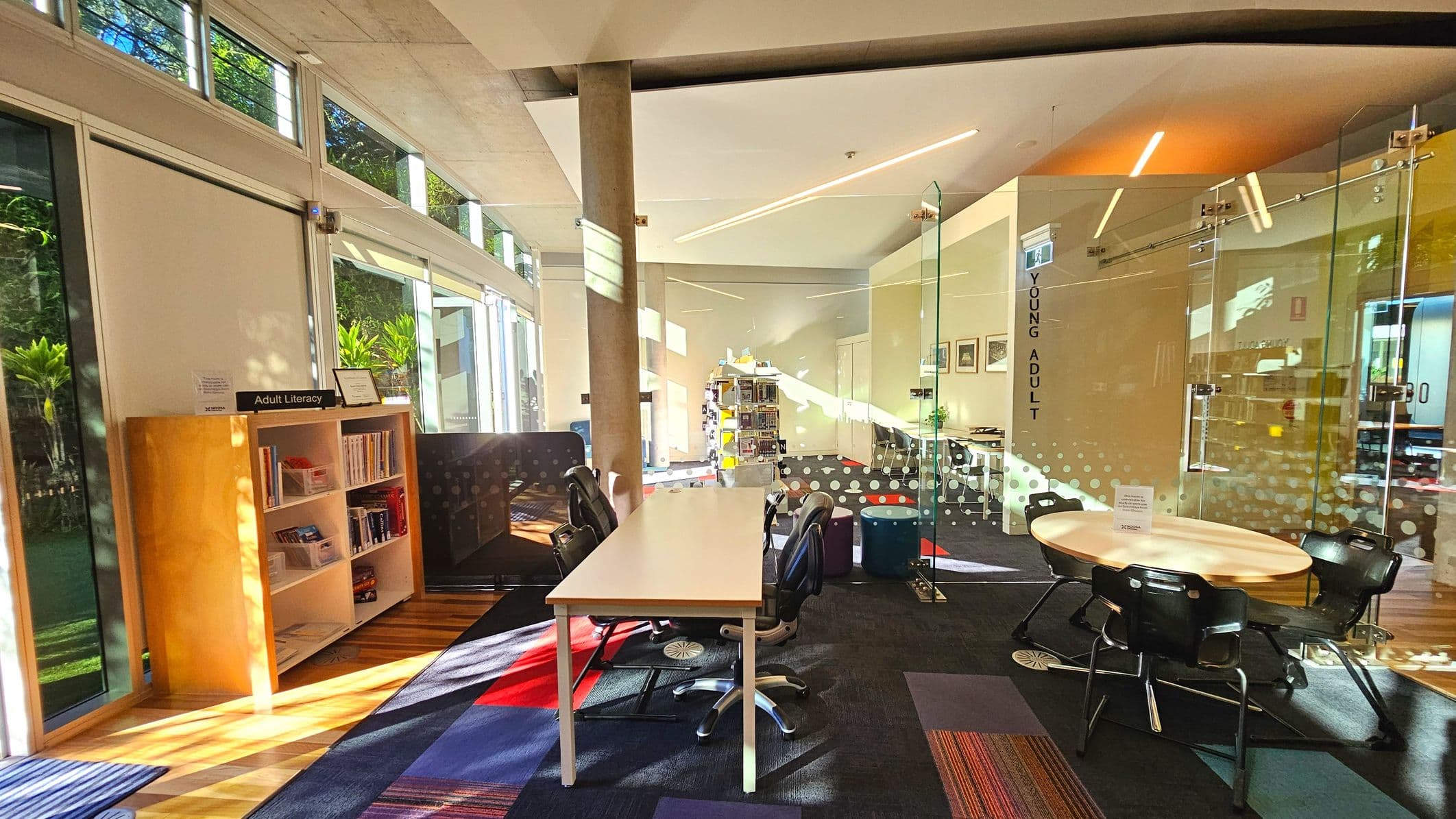 A library interior featuring a modern design with large glass windows and a glass-walled room labelled 'Young Adult'. A wooden bookshelf marked 'Adult Literacy' is filled with books. A long white table with office chairs is centrally placed on a carpeted floor with colourful patterns. A round table with chairs is nearby. The space is well-lit with natural light streaming through the windows, highlighting the open and airy layout.