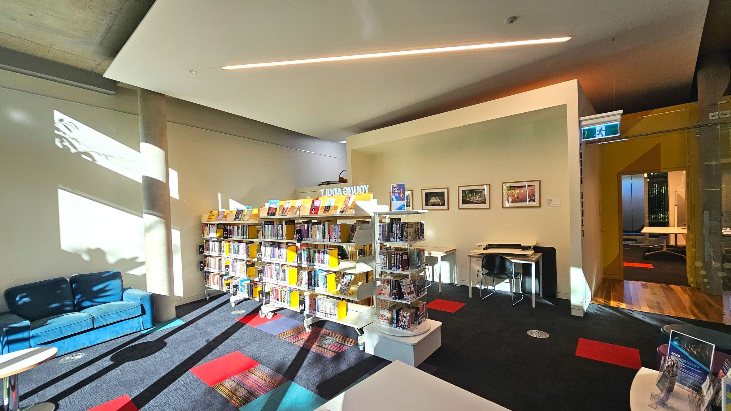 A library interior with a focus on a 'Young Adult' section featuring two white bookcases filled with books, positioned on a dark carpet with red and multicoloured square patterns. To the left, a blue sofa sits against a wall with light patterns. On the right, a small desk with a chair is visible, and a glass door leads to another room with wooden flooring. The ceiling has a modern light fixture, and framed pictures adorn the wall.