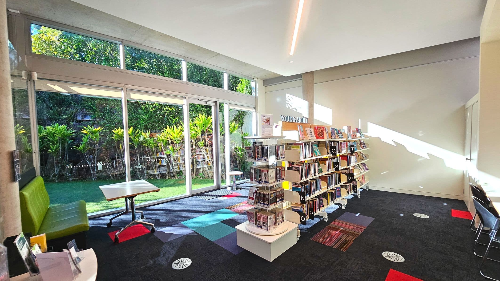 A library room with large glass sliding doors on the left, revealing a view of lush greenery outside. Inside, there are bookshelves labelled 'Young Adult' filled with books, arranged in rows. A green cushioned bench and a small table with wheels are near the doors. The floor is carpeted with a mix of dark and colourful square patterns. The space is well-lit with natural light streaming through the windows.
