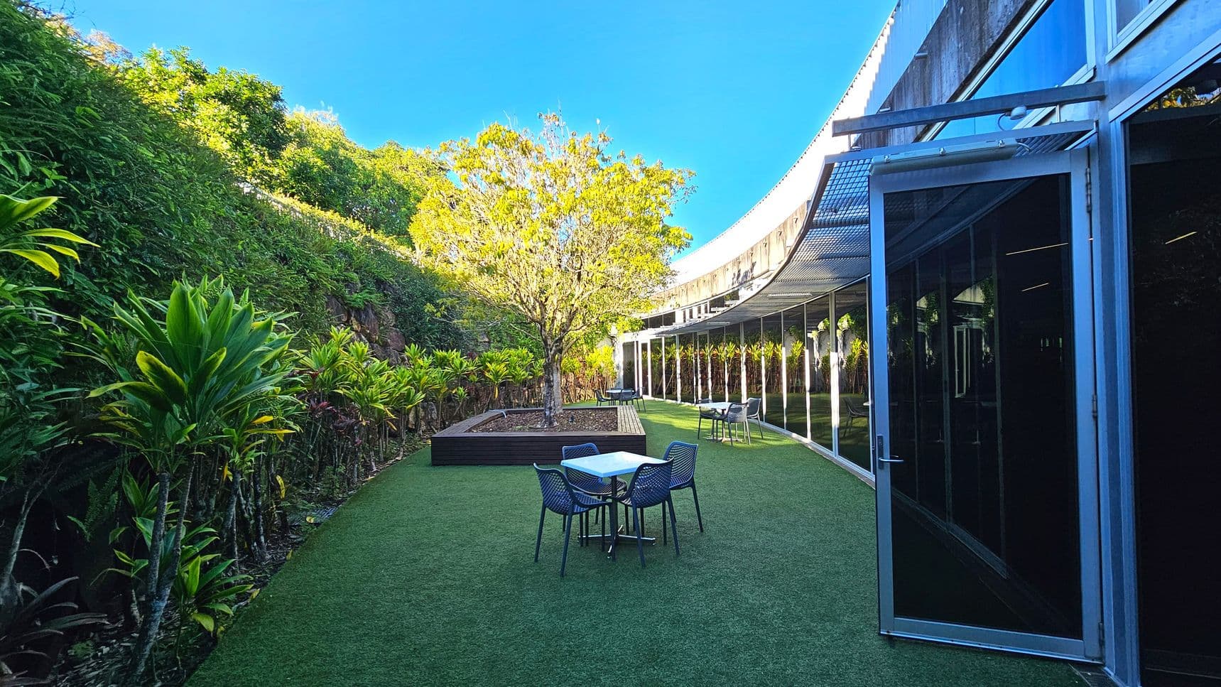 An outdoor area with artificial grass flooring features a central tree in a raised planter. Surrounding the space are lush green plants along a sloped embankment. On the right, a curved glass building with open doors reflects the greenery. Several tables with chairs are scattered across the grass, suggesting a seating area. The space feels open, bordered by the building and the embankment.