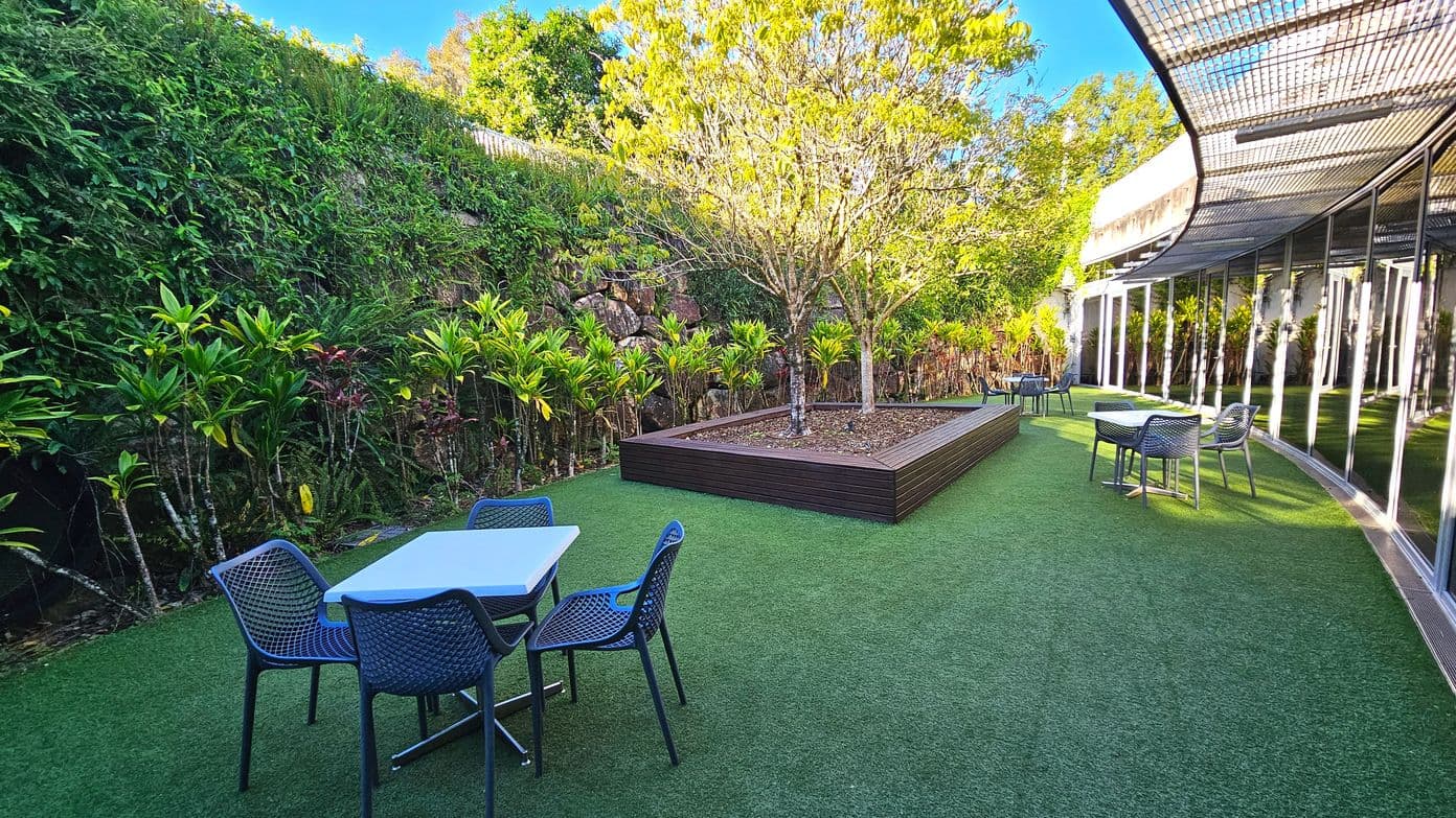 An outdoor area with artificial grass flooring features several blue mesh chairs and tables. A raised wooden planter box with a tree is centrally located. To the left, a lush green vertical garden covers a stone wall. On the right, a building with large reflective glass windows and a curved metal awning provides shade. The space is open and bordered by greenery, creating a tranquil setting.