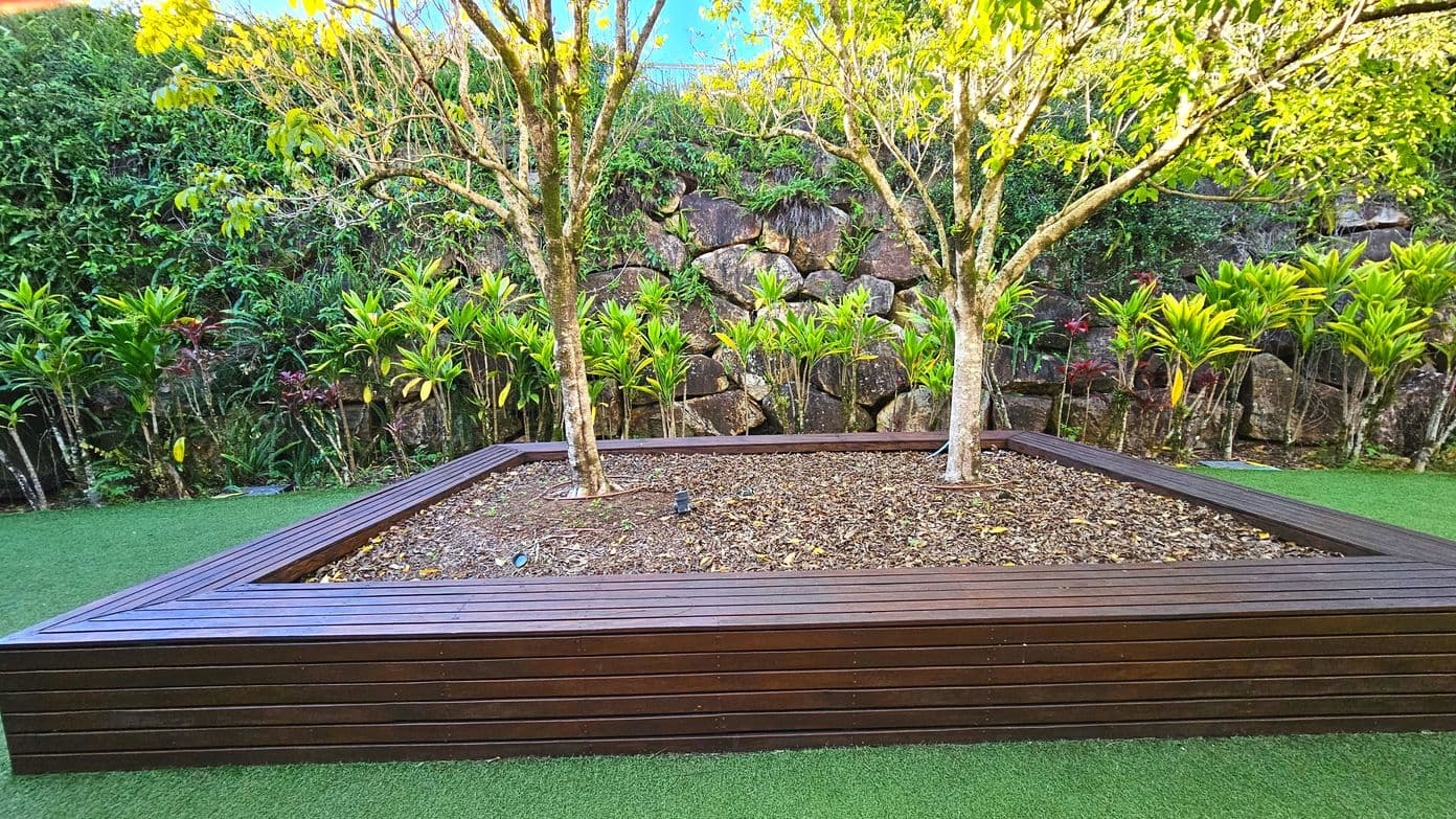 A landscaped garden area featuring a large, raised wooden planter box with two trees growing from it. The planter is rectangular and made of dark wood slats, filled with mulch. Surrounding the planter is a neatly trimmed artificial grass surface. In the background, a stone retaining wall is partially covered with lush green foliage and small shrubs, creating a natural boundary.