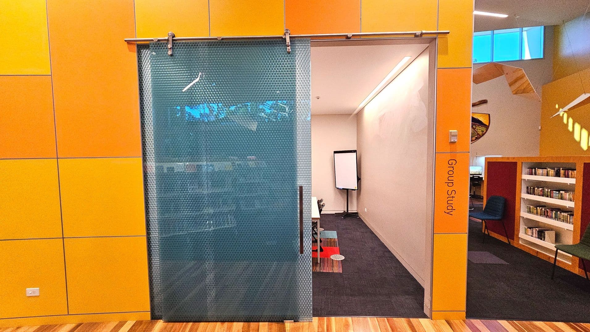 A room labelled 'Group Study' with a partially open sliding door made of textured glass. The walls are covered in large orange panels. Inside, there is a dark carpeted floor with a colourful rug, a whiteboard on a stand, and a table with chairs. Adjacent to the room, there is a small library area with shelves of books and a green chair. The ceiling has a modern design with angular features and a high window.