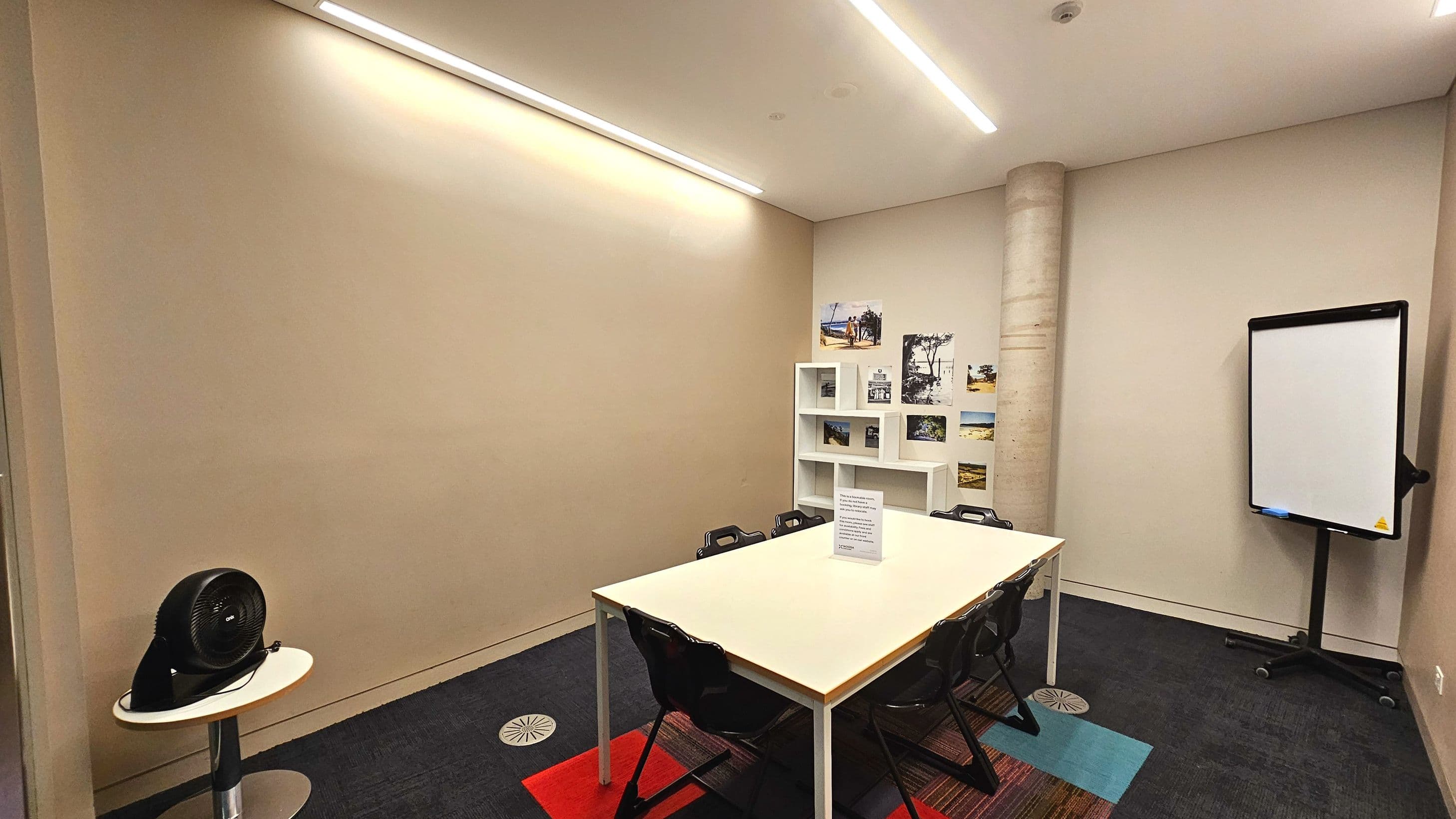 A small meeting room with a rectangular white table surrounded by black chairs on a dark carpet with colourful square patterns. A cylindrical concrete pillar stands in the corner near a white shelving unit displaying photos. A digital whiteboard on wheels is positioned against the wall. A small round table with a fan is in the foreground. The room is enclosed with light-coloured walls and ceiling lights.