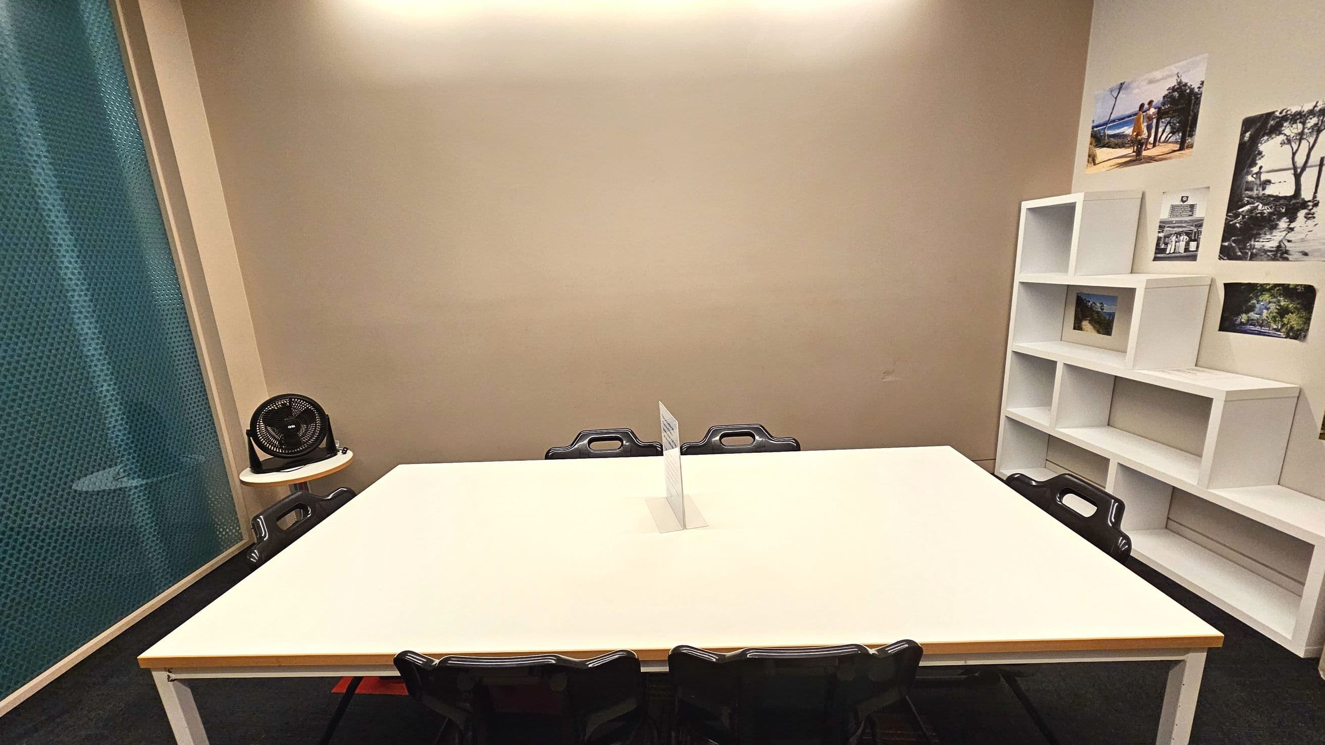 A small room with a large white rectangular table at the centre, surrounded by black chairs. A small fan sits on a round side table to the left. The back wall is plain, while the right wall features a white stepped shelving unit with various photographs displayed above it. The floor is carpeted in dark grey, and a blue patterned curtain is partially visible on the left side.