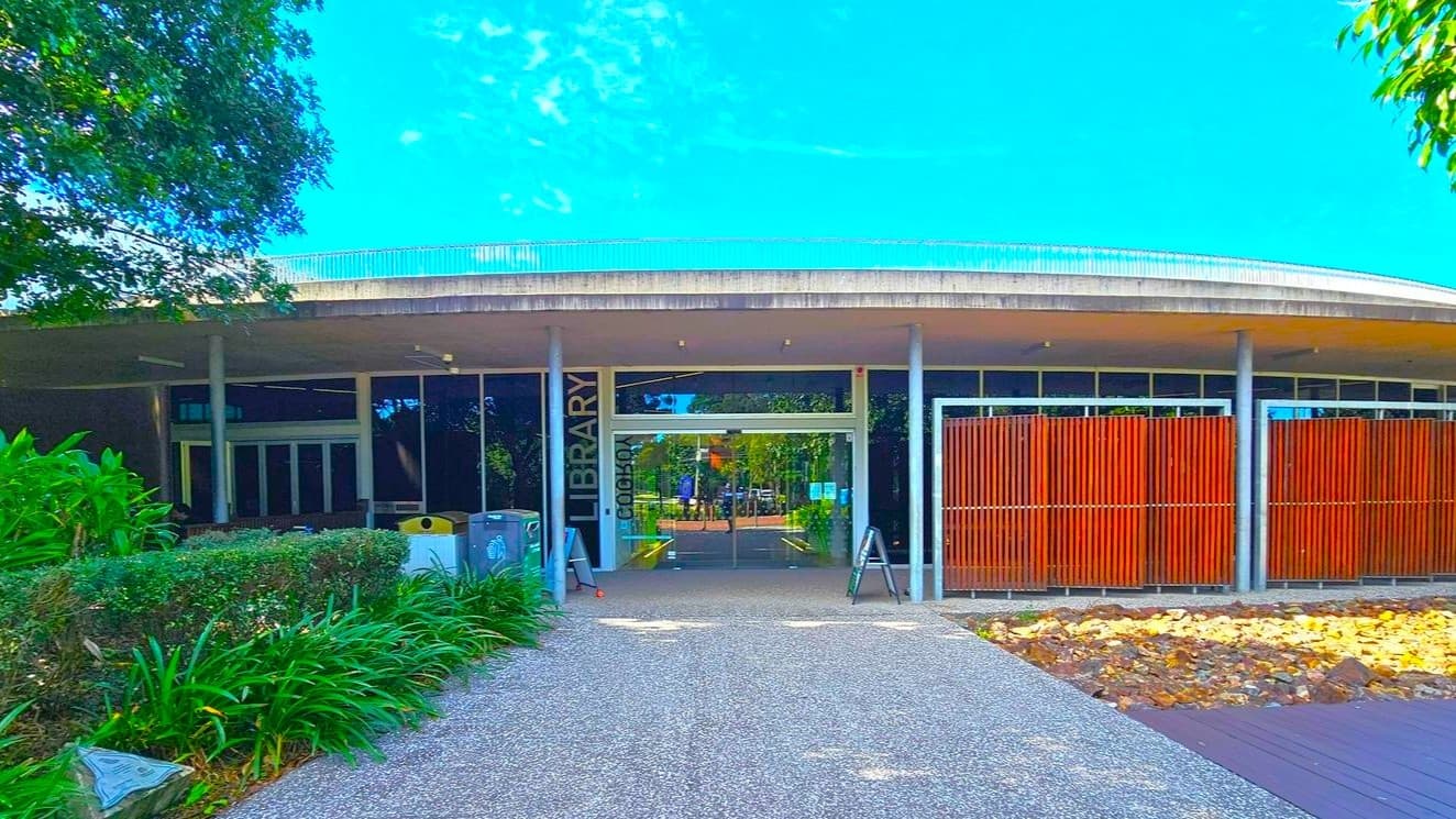 A building entrance is flanked by lush green trees and shrubs. The path leading to the entrance is bordered by bollards and a red hydrant box marked 'HYDRANT BOOSTER' is visible on the left. The building has a flat roof and large glass windows. Parking spaces are marked on the asphalt in the foreground. Signs are posted near the entrance, and a bicycle rack is partially visible on the left.