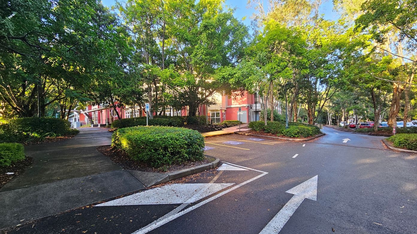 A paved road with white directional arrows leads towards a red building partially obscured by lush green trees and shrubs. The road splits into two paths, with one leading to a parking area marked with blue disabled parking signs. Concrete pavements run alongside the road, bordered by manicured hedges. Tall street lamps are visible, and several parked cars can be seen in the distance.
