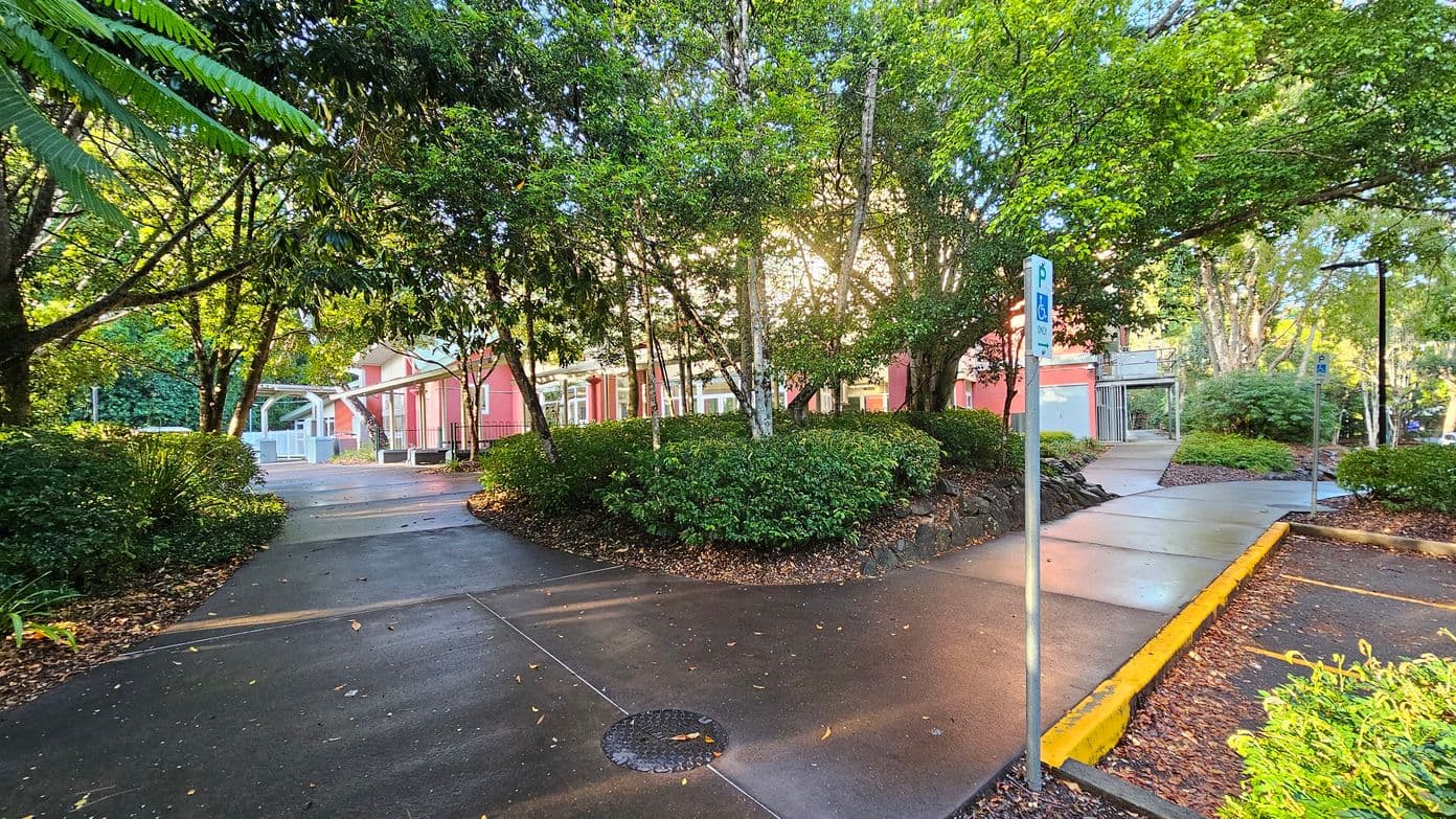 A paved pathway curves through a lush garden area with dense green foliage and tall trees. The path splits, leading towards a red building with large windows and a covered entrance. Parking signs for disabled access are visible along the path. The area is well-shaded by the trees, and the ground is wet, suggesting recent rain. A yellow kerb marks the edge of the path, and a metal grate is embedded in the pavement.