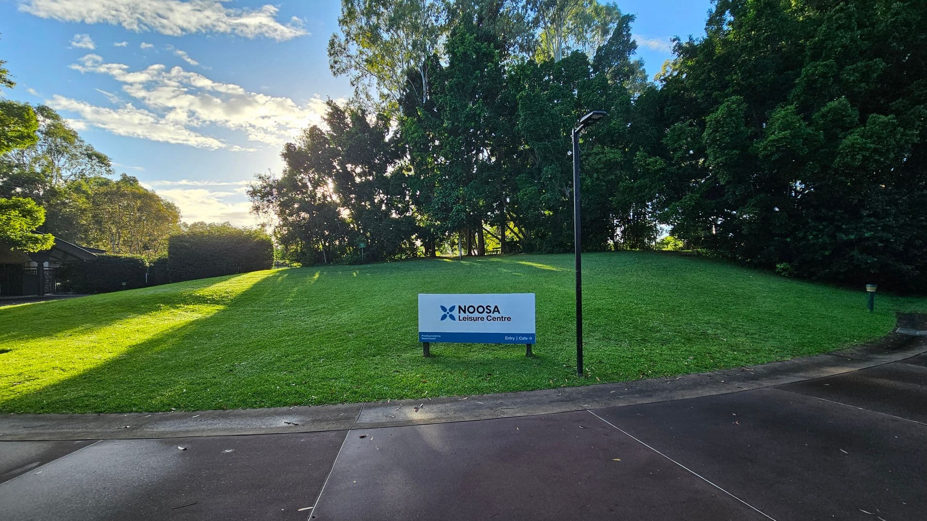 A gently sloping grassy hill is bordered by dense trees on the right and a hedge on the left. In the foreground, a concrete path curves slightly, leading towards a white sign that reads 'Noosa Leisure Centre'. A tall, slender lamp post stands near the sign. The area feels open, with the grass and path providing a clear transition between surfaces.