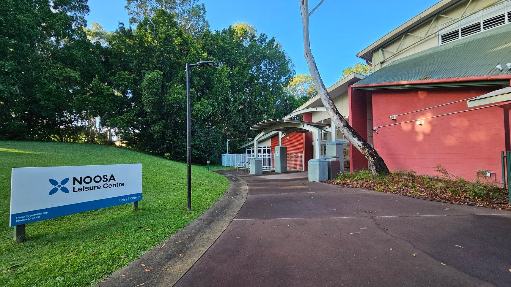A pathway curves gently towards the entrance of a building with a red exterior wall and a grey corrugated roof. A large sign on the left reads 'Noosa Leisure Centre' with directions to the entry and café. The path is bordered by a grassy area and a row of trees. A streetlamp stands beside the path, and there are bike racks and bins near the entrance, which is covered by a metal awning.