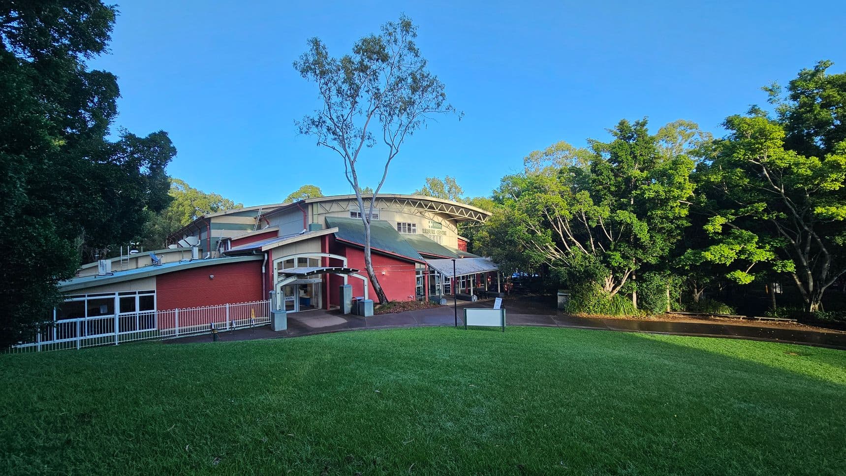 A red brick building with a green roof and a prominent arched entrance is situated on a grassy slope. The structure features large windows and a white fence along one side. A tall tree stands in front, and dense greenery surrounds the area, creating a natural boundary. A paved path leads to the entrance, and a sign is visible near the path. The building appears to be a community or leisure centre.
