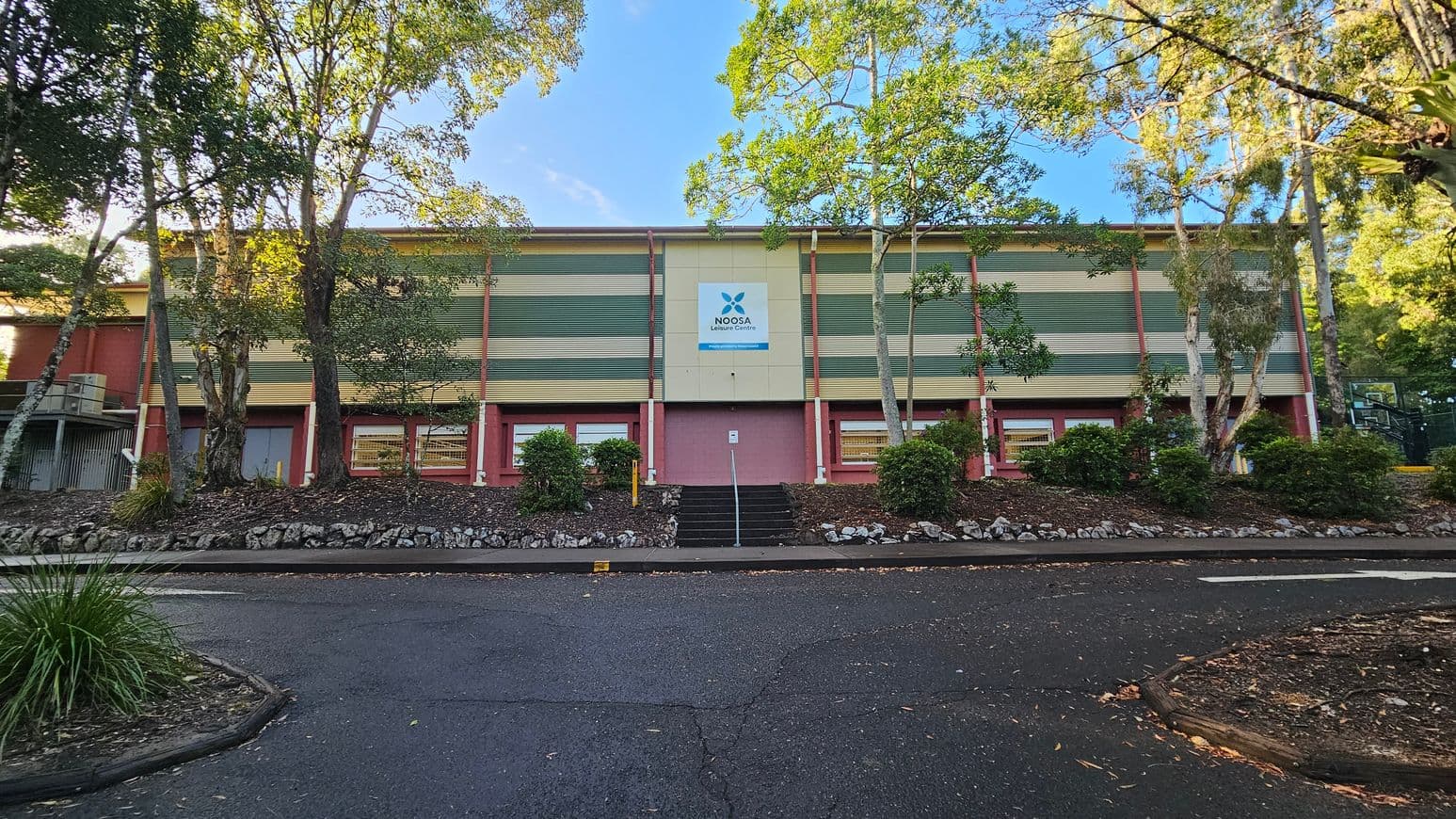 A rectangular building with a sign reading 'Noosa Leisure Centre' is visible, featuring a facade of horizontal green and beige panels. The structure is elevated with a central staircase leading up to it, flanked by small shrubs and trees. The area in front is paved with asphalt, bordered by a low stone wall and additional greenery. The building is surrounded by tall trees, creating a semi-enclosed environment.