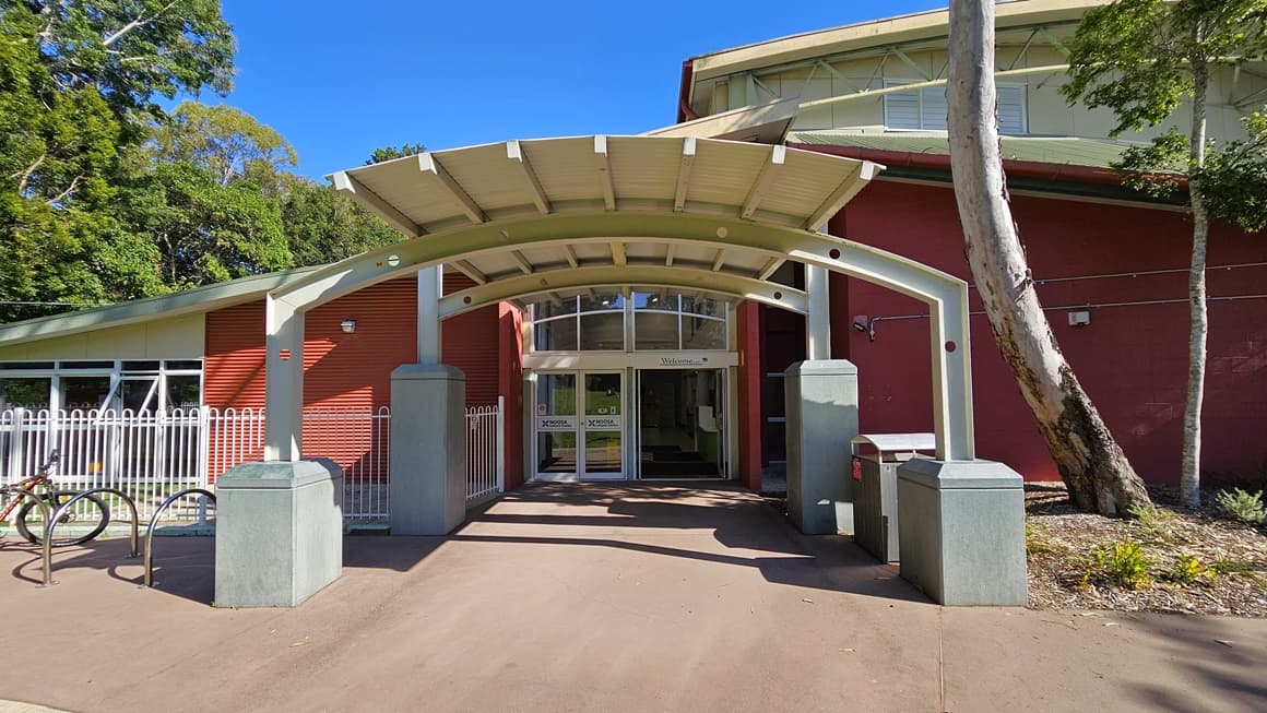 A building entrance with a prominent arched metal canopy supported by white pillars. The facade is red with large windows above the glass double doors. A white metal fence runs along the left side, with a bicycle rack nearby. The path leading to the entrance is paved, and there are trees and shrubs on the right. The structure appears modern and functional, with clear access to the interior.