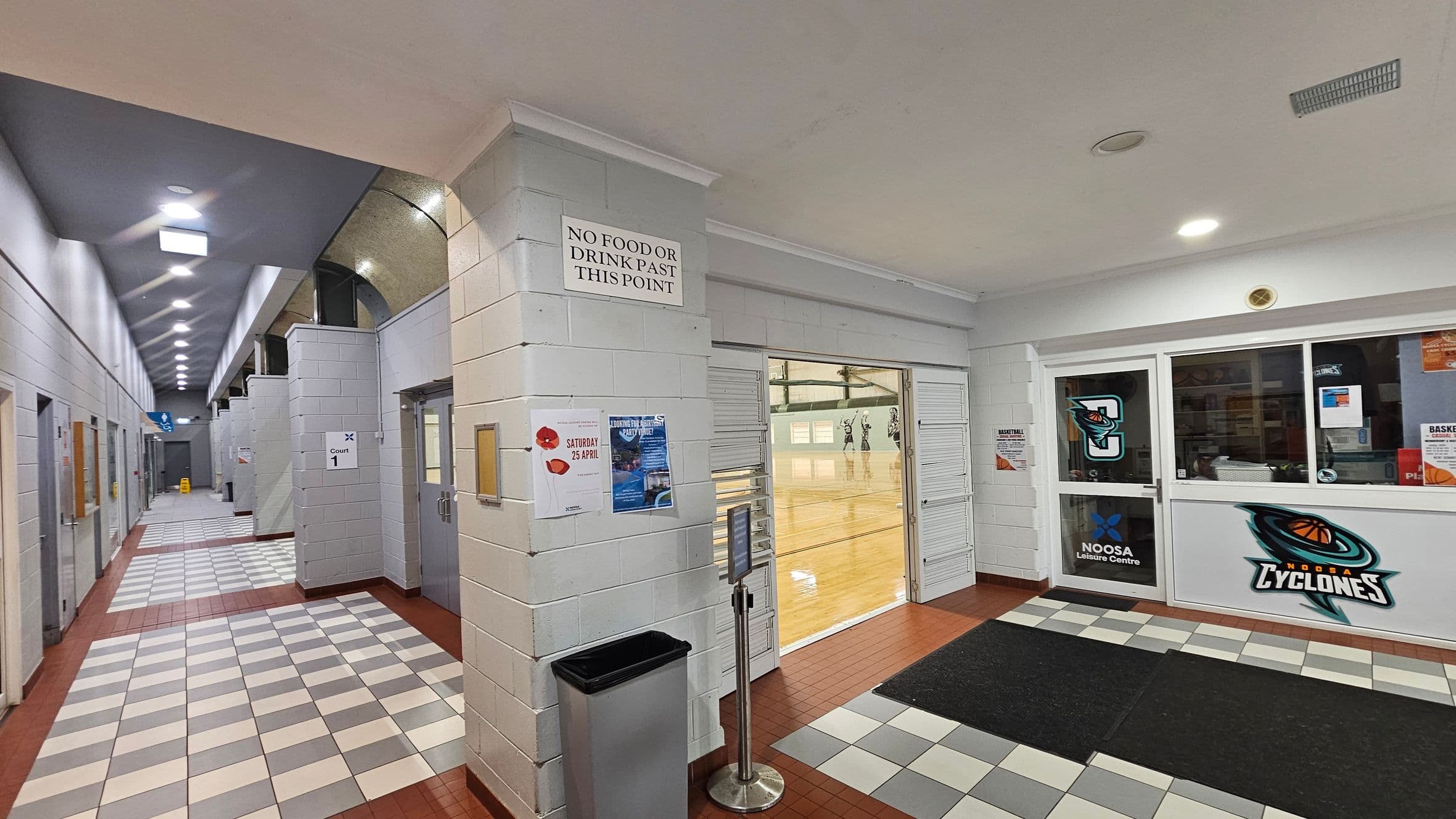 A corridor with white brick walls and a tiled floor featuring a pattern of white and grey squares bordered by red tiles. On the right, a glass door with 'Noosa Cyclones' signage leads to a sports court visible through a window. A sign above reads 'No food or drink past this point'. The corridor extends left with multiple doors and overhead lighting. Posters and a bin are mounted on the walls.