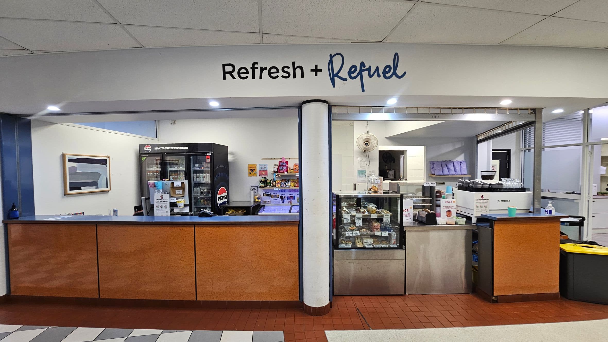 A small refreshment kiosk with a sign reading 'Refresh + Refuel' above. The counter is divided into two sections with a central column. On the left, a black Pepsi fridge is stocked with drinks, and shelves display snacks. On the right, a coffee machine and a glass display case with food items are visible. The floor transitions from red tiles in front of the counter to a patterned surface in the foreground.