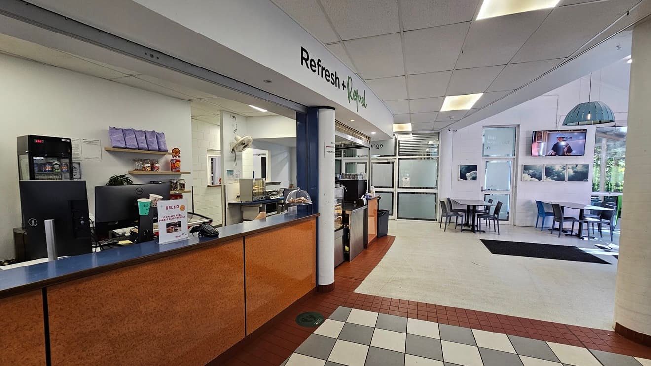The image shows a cafeteria area with a counter on the left featuring a blue countertop and wooden panels. Behind the counter are shelves with items and a small refrigerator. A sign above reads 'Refresh'. To the right, there are tables and chairs arranged in a seating area with large windows and wall-mounted screens. The floor transitions from red tiles near the counter to white tiles in the seating area.