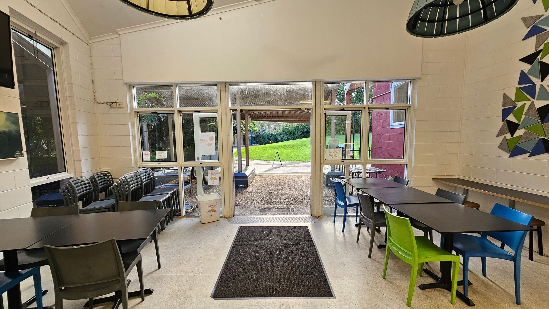 A room with a high ceiling features a large glass door leading outside to a grassy area. Inside, there are several black tables with colourful chairs, including green and blue. Stacked chairs are visible on the left. The floor is light-coloured with a dark mat near the entrance. A geometric wall decoration is on the right. The space feels open with large windows and a view of the outdoors.