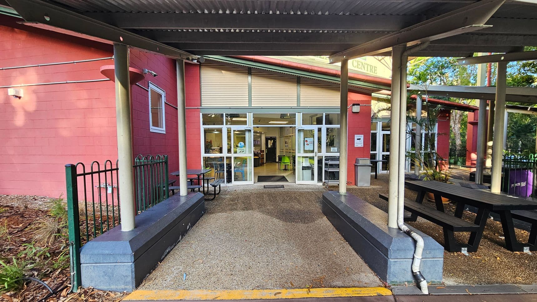 The image shows the entrance to a building with a red brick facade and a large glass door framed by white panels. A corrugated metal awning supported by grey pillars extends over the entrance. To the left, a black metal fence borders a garden area. Picnic tables with benches are positioned on either side of the entrance. The ground is covered with a textured concrete surface, transitioning to a smooth interior floor visible through the glass doors.