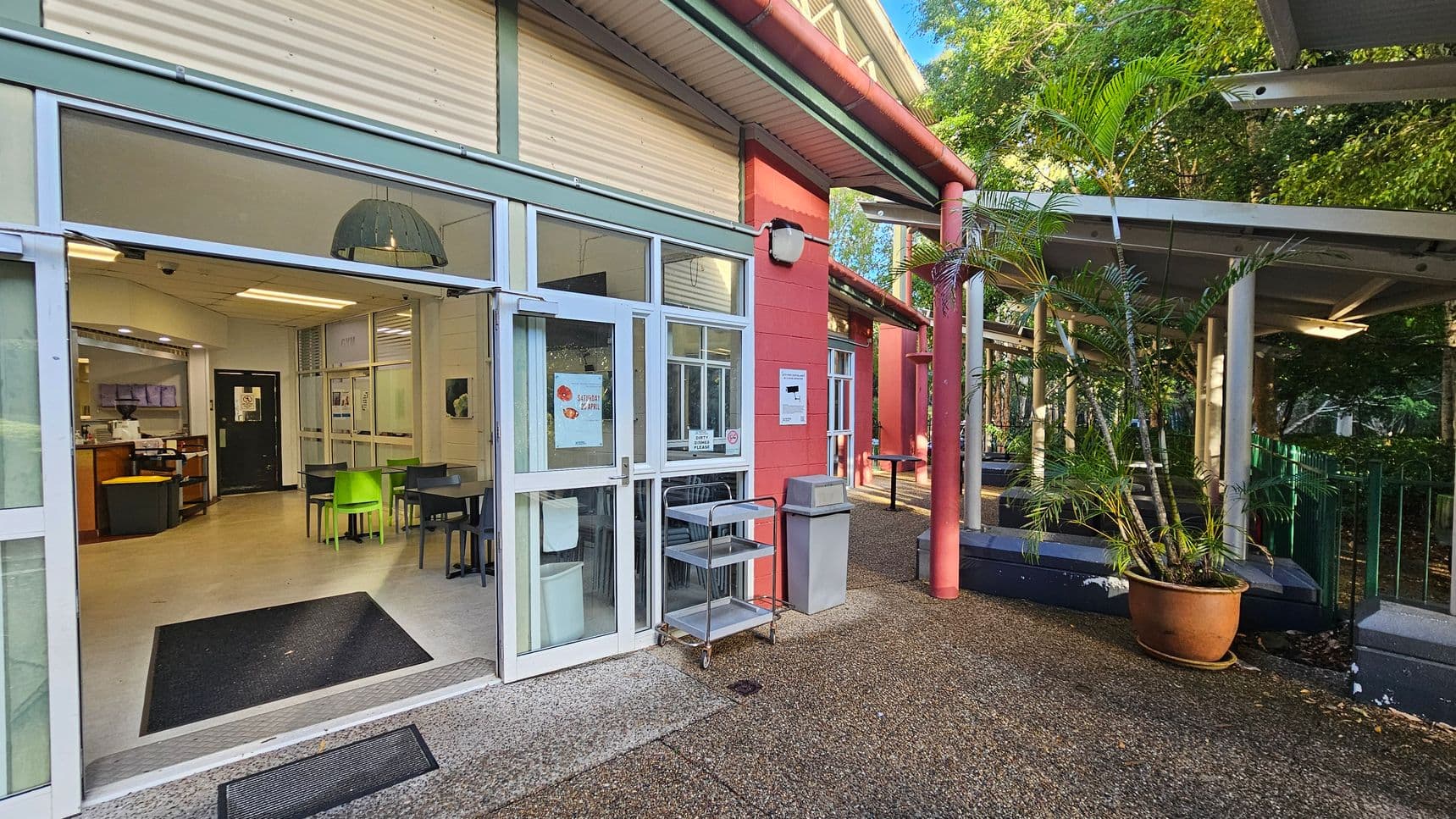 The image shows the entrance to a building with large glass doors and windows framed in white. Inside, there are tables and chairs, some in bright green. A black mat lies at the threshold. Outside, a red wall with a sign and a grey bin are visible. A metal trolley stands near the entrance. A covered walkway with potted plants and a textured ground surface extends to the right.