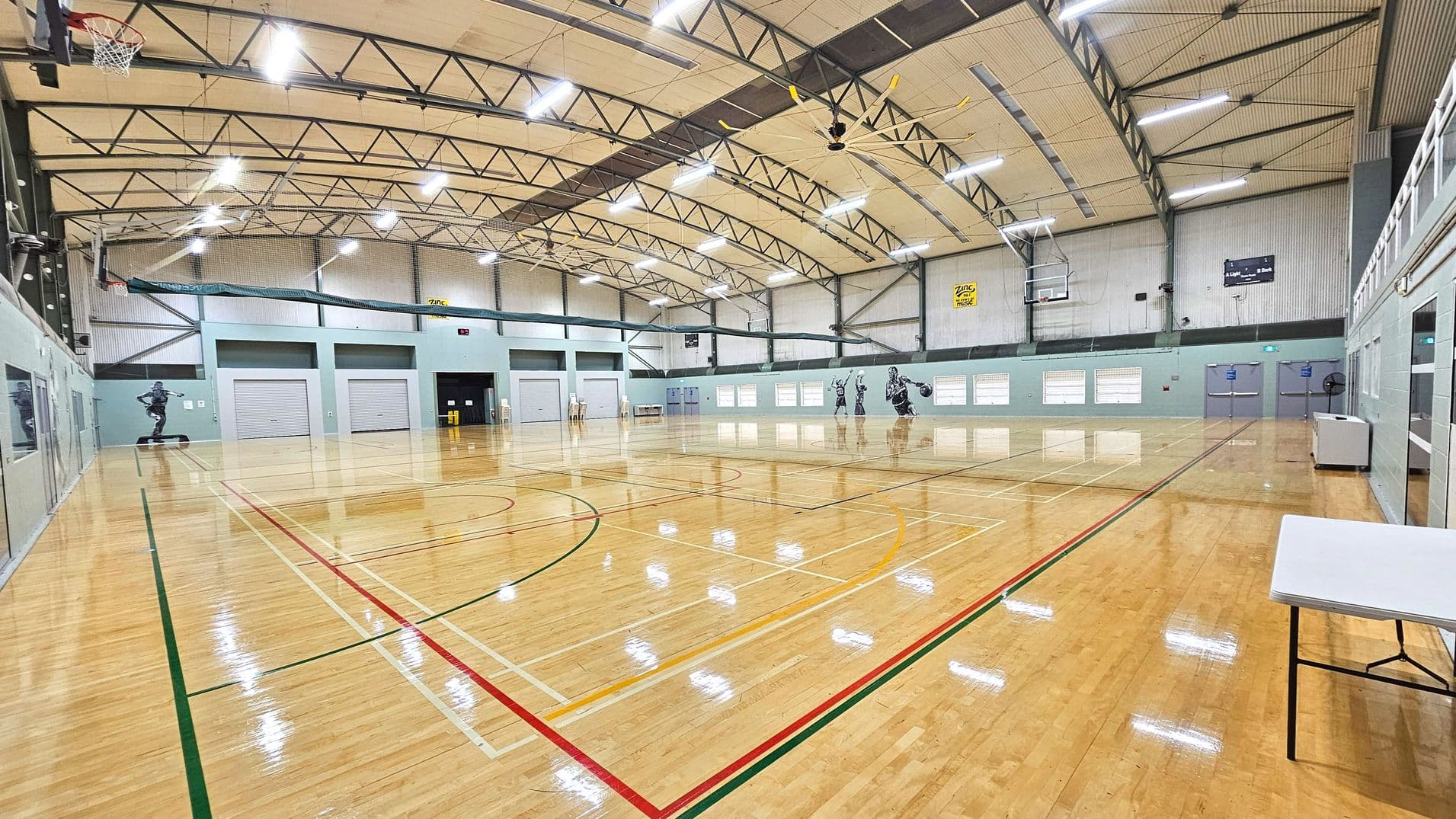 A spacious indoor sports hall with a polished wooden floor marked with various sports lines in red, green, and yellow. The ceiling is high with exposed metal beams and bright overhead lights. On the left, a basketball hoop is mounted, and the walls feature large murals of athletes. Several closed roller doors line the far wall, and a white folding table is positioned on the right. The space feels open and expansive.