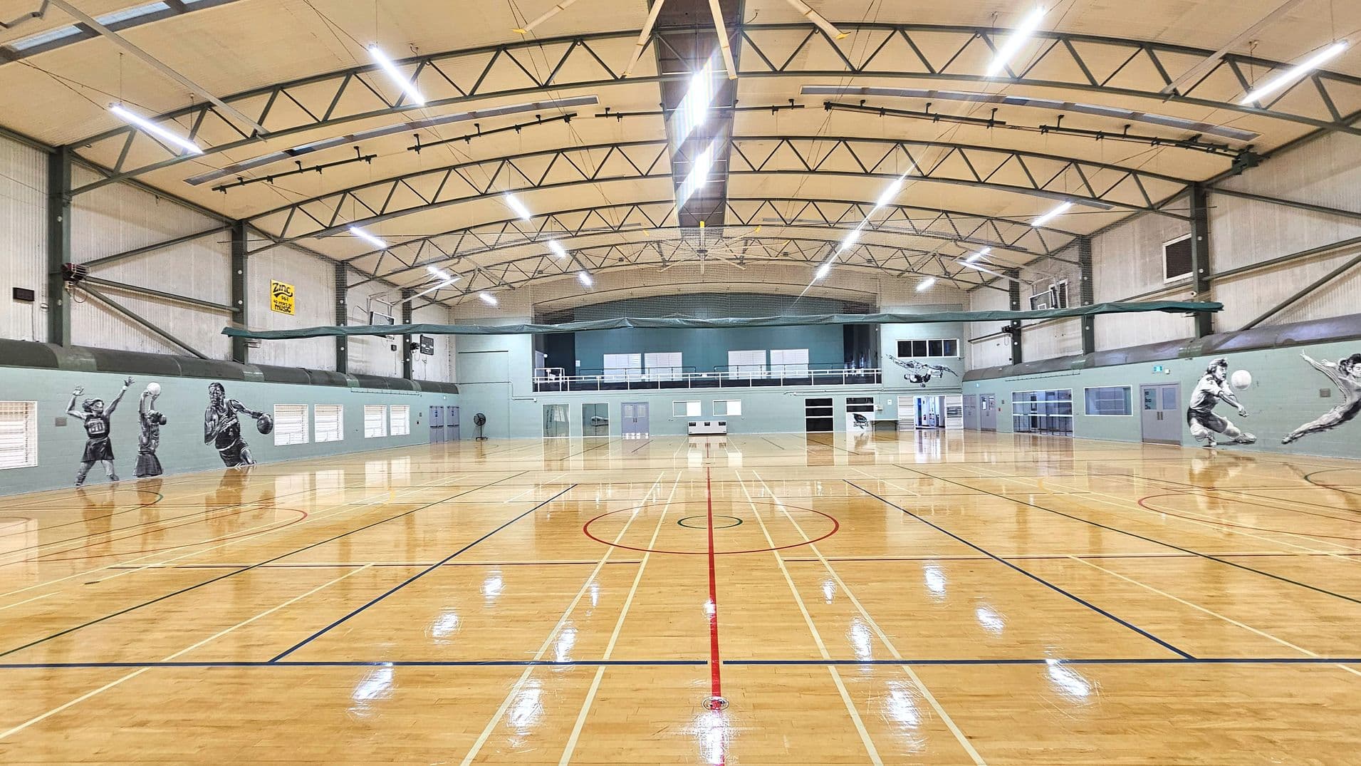 A spacious indoor sports hall with a polished wooden floor marked for various sports. The ceiling features exposed metal beams and bright overhead lighting. The walls are adorned with large murals of athletes in action. A mezzanine level with windows and doors is visible at the far end. The hall is enclosed by light-coloured walls with multiple doors and windows along the sides.