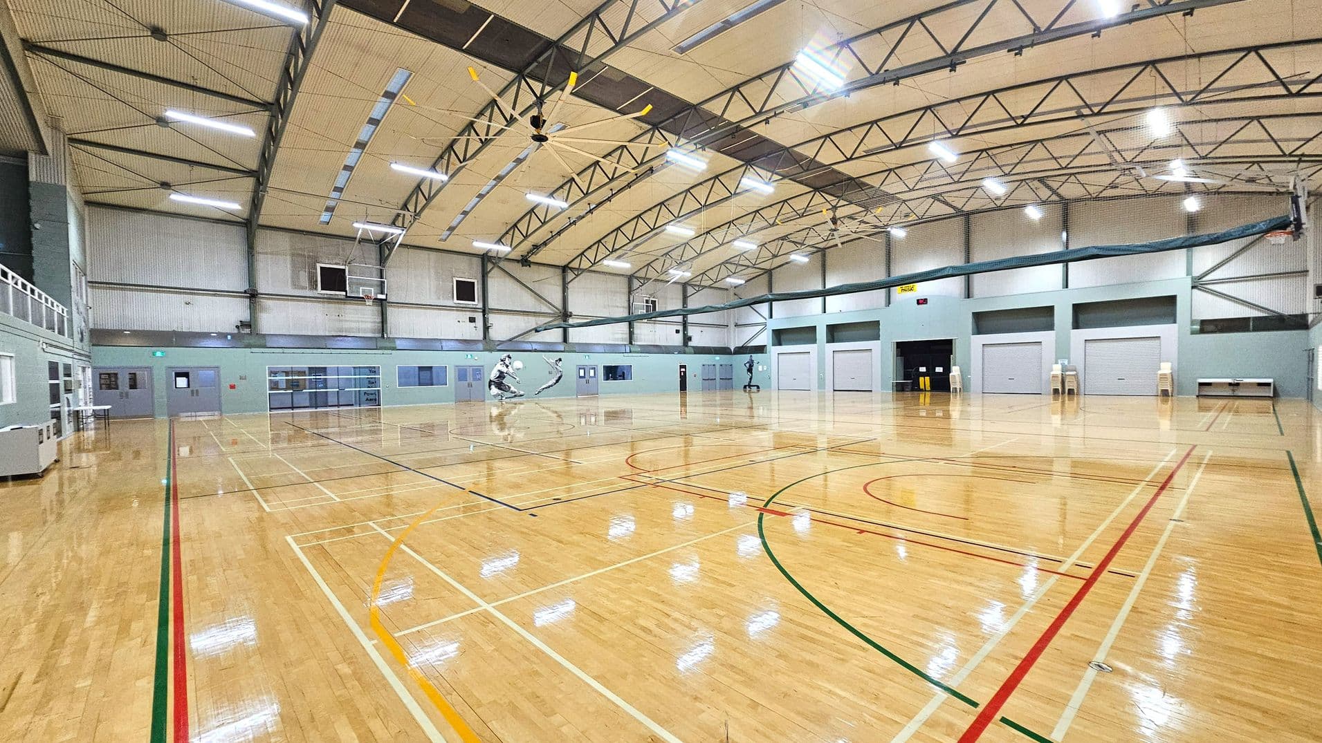 A spacious indoor sports hall with a polished wooden floor marked with various sports lines in red, green, and yellow. The ceiling is high with exposed metal beams and bright overhead lights. On the far wall, there are multiple closed roller doors and a scoreboard. Basketball hoops are mounted on the walls. The space is enclosed by light blue walls with several doors and windows.