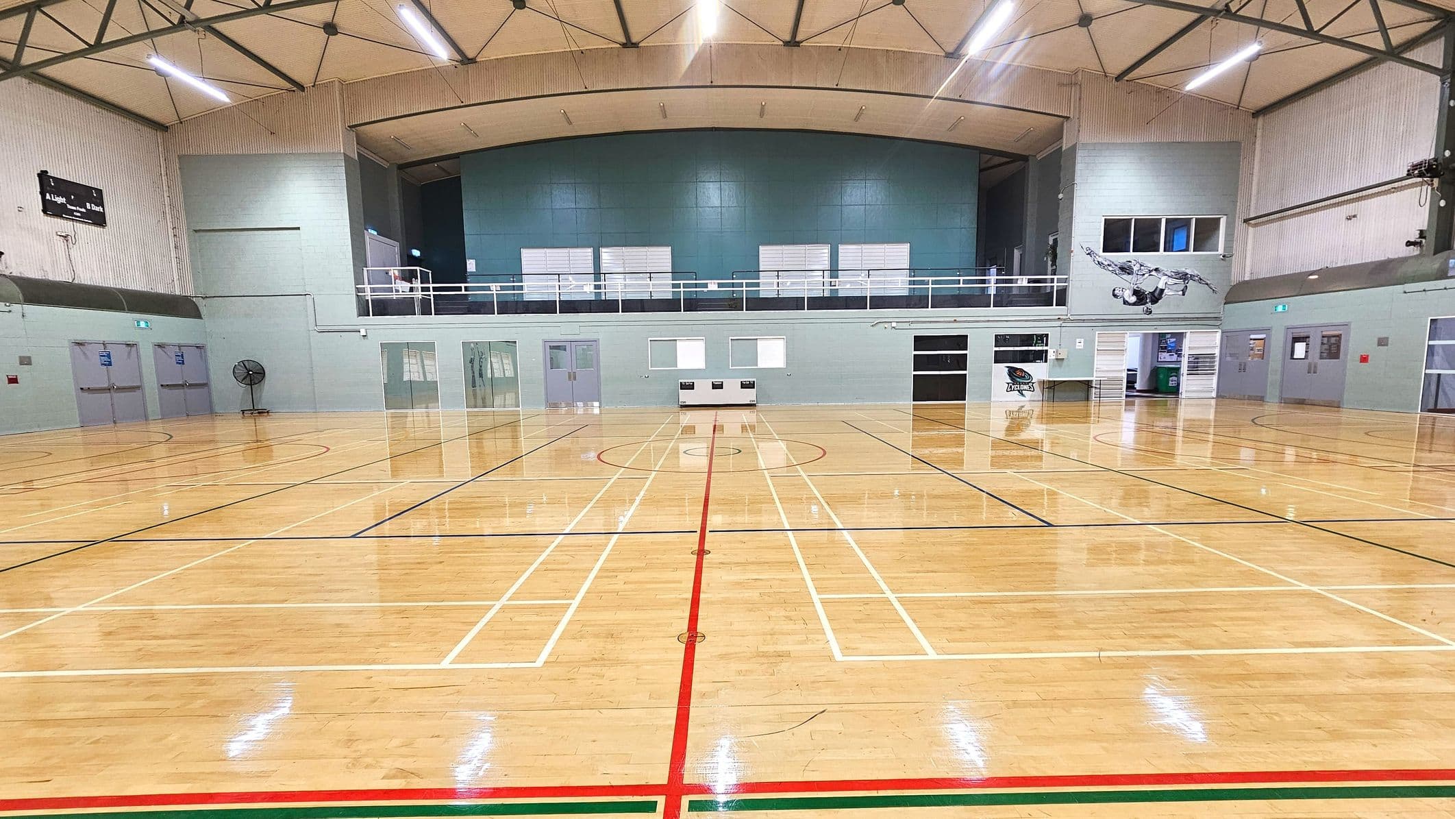 A spacious indoor sports hall with a polished wooden floor marked for various sports. The walls are light grey with a large mural of an athlete on the right. A scoreboard is mounted high on the left wall. The ceiling is high with exposed beams and fluorescent lighting. A mezzanine level with railings runs along the back wall, above several doors and windows. A large fan is positioned near the left wall.