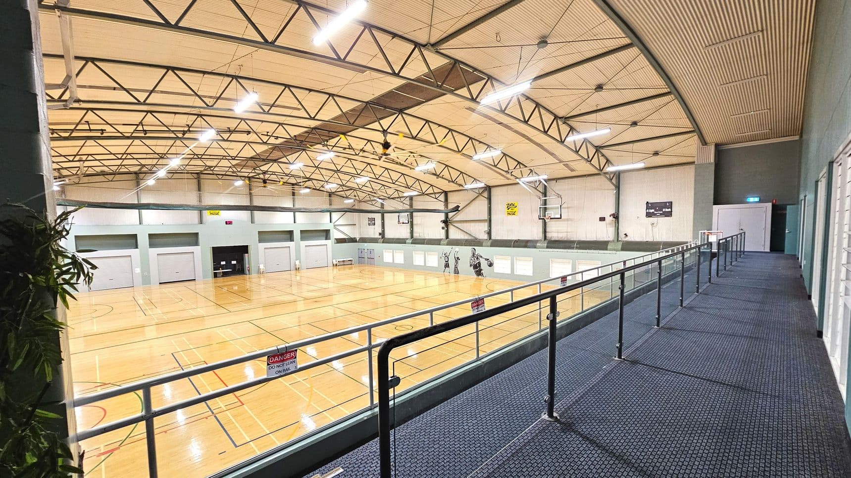 An indoor sports hall with a high arched ceiling supported by metal trusses. The floor is a polished wooden surface marked for basketball. A mezzanine level with a blue carpeted walkway and metal railing overlooks the court. Signs on the railing warn against leaning. The walls are grey with large closed shutters and sports-themed murals. Emergency exit signs and doors are visible along the walkway.