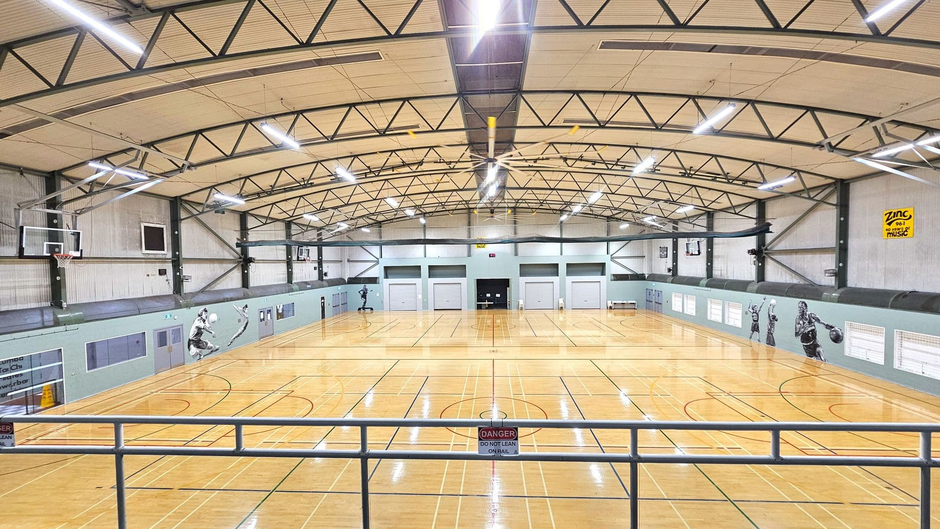 A spacious indoor sports hall with a high arched ceiling supported by metal beams. The polished wooden floor is marked with various sports lines. On the left and right walls, there are basketball hoops and large sports-themed murals. The far wall features multiple roller doors and a central entrance. Fluorescent lights hang from the ceiling, and a metal railing with a warning sign is visible in the foreground.