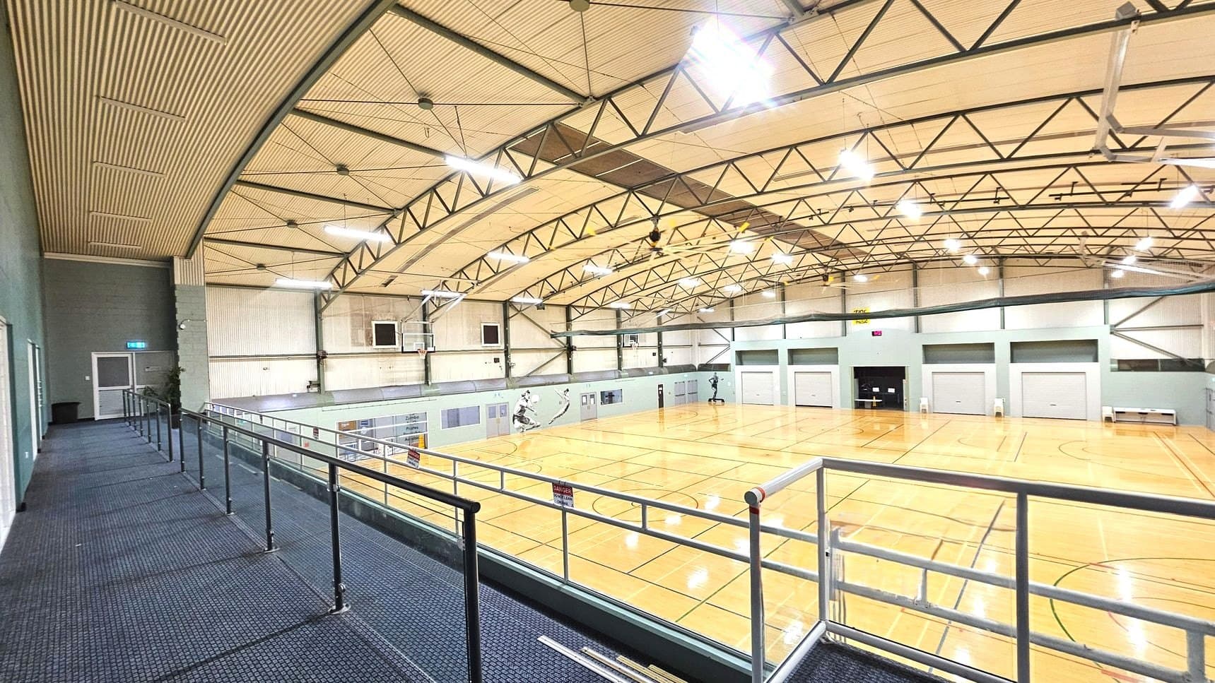 A spacious indoor sports hall with a high, arched ceiling supported by metal trusses. The floor is polished wood with basketball court markings. A mezzanine level with a blue carpeted walkway and metal railing overlooks the court. Several closed roller doors line the far wall. Fluorescent lights illuminate the space, and a basketball hoop is visible on the left wall.